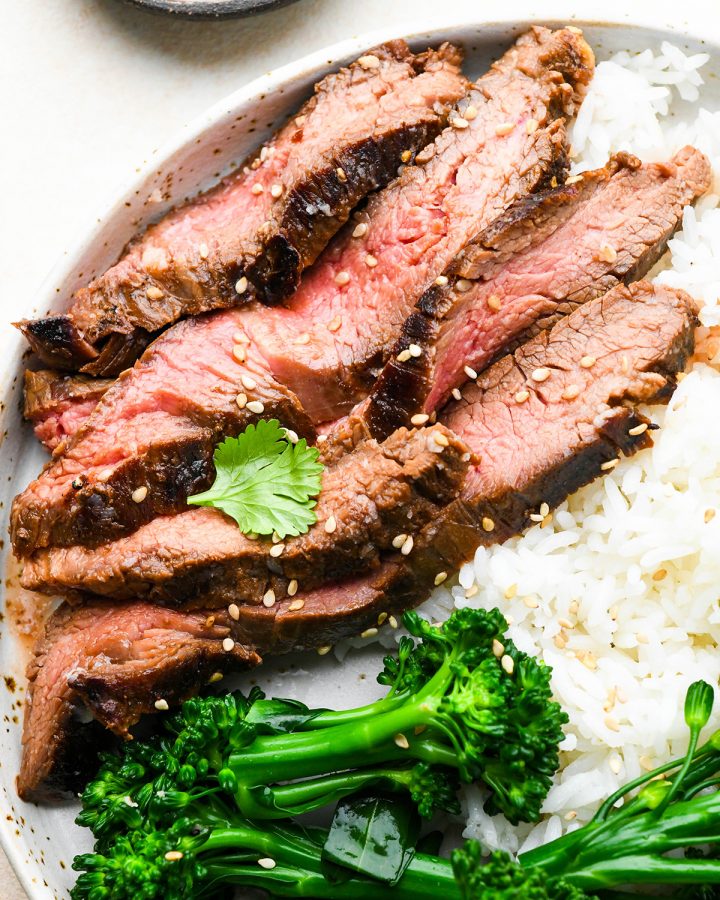4 slices of grilled flank steak after soaking in an asian flank steak marinade on a plate with rice and broccoli