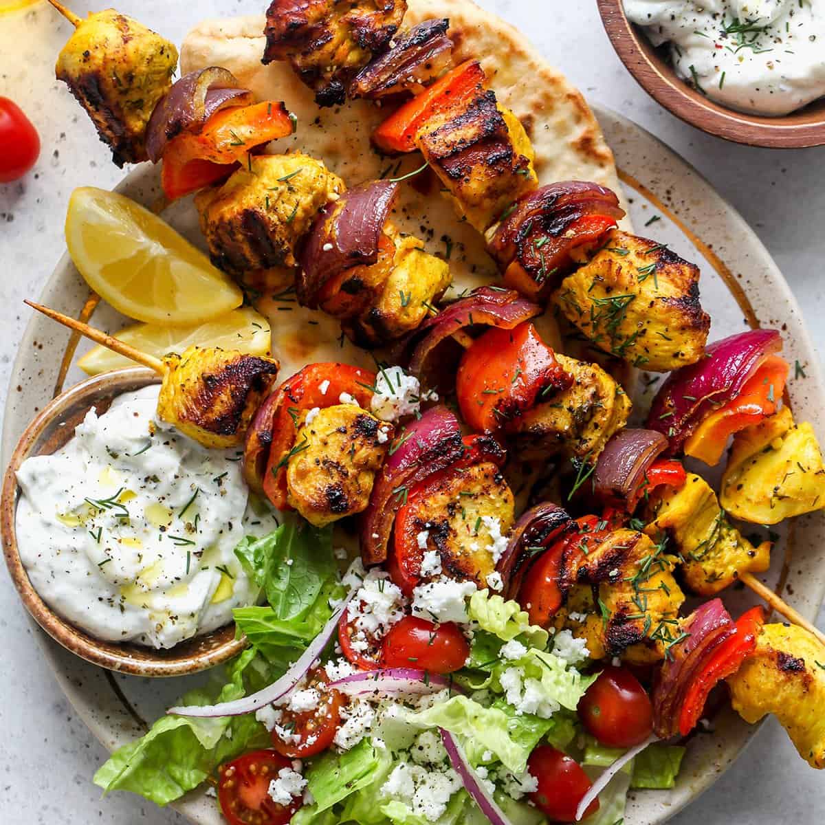 3 chicken kebabs on a plate with a salad and tzatziki