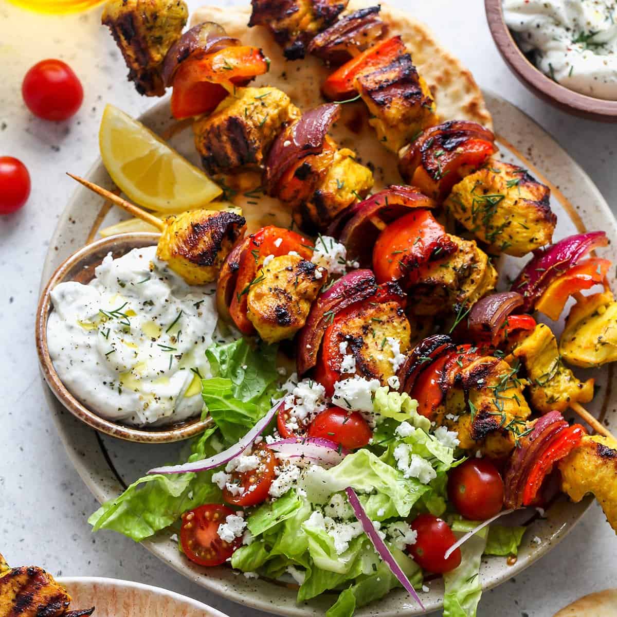 3 chicken kabobs on a plate with a salad and tzatziki