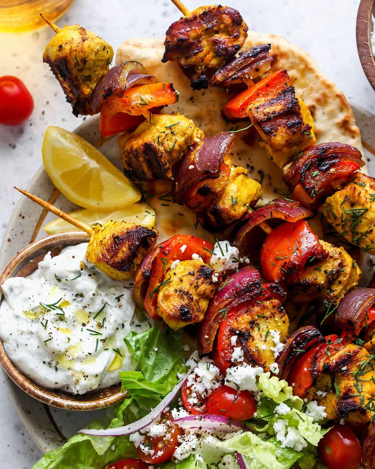 3 chicken kabobs on a plate with a salad and tzatziki