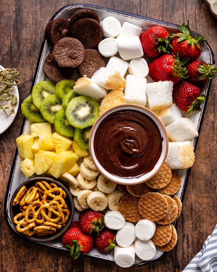 a bowl of Chocolate Fondue surrounded by fruit, cake, brownies, cookies and pretzels