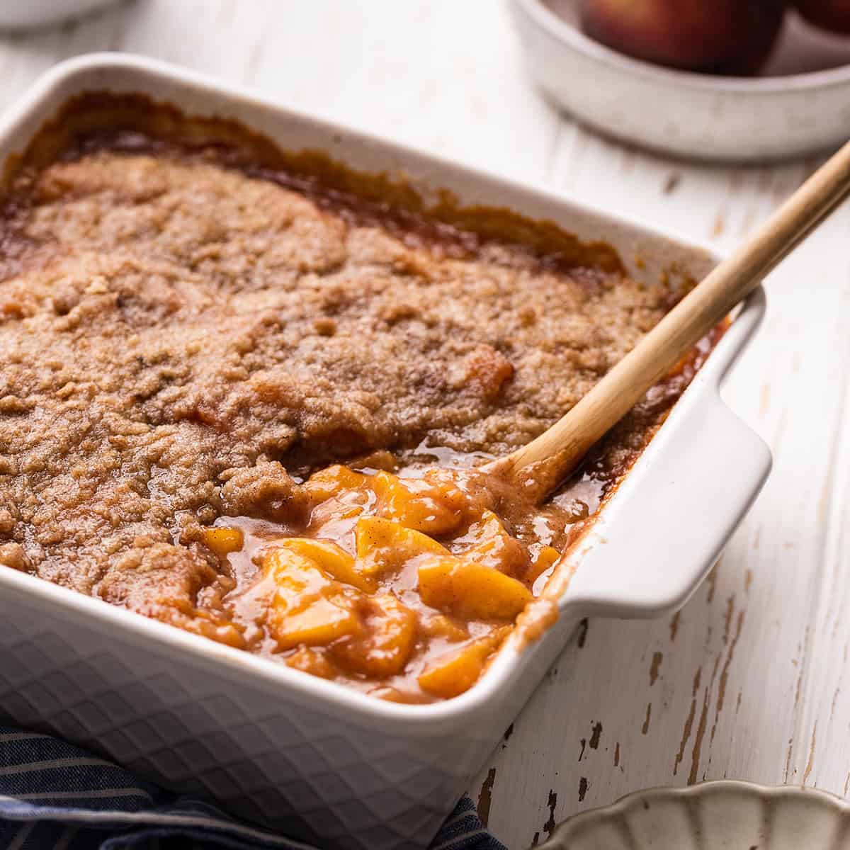 Peach Crumble in a baking dish with a wooden spoon serving a portion