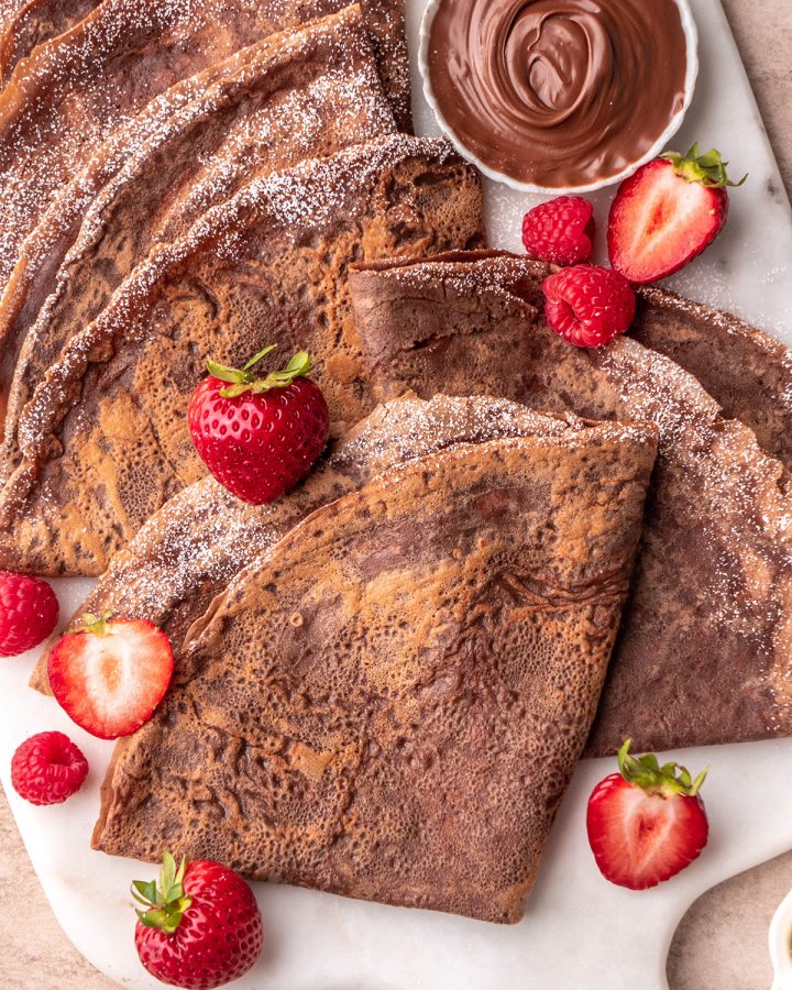 5 Chocolate Crepes folded into triangles on a plate with berries and powdered sugar on a serving platter