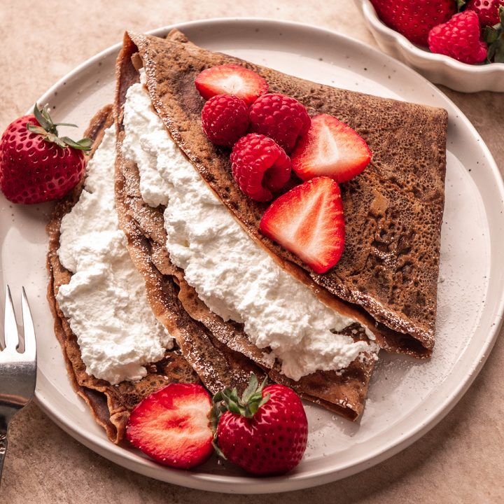 two Chocolate Crepes on a plate filled with whipped cream and topped with berries