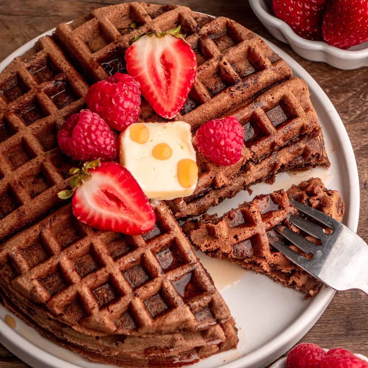 Chocolate Waffles on a plate with butter, berries and syrup and a fork taking a bite 