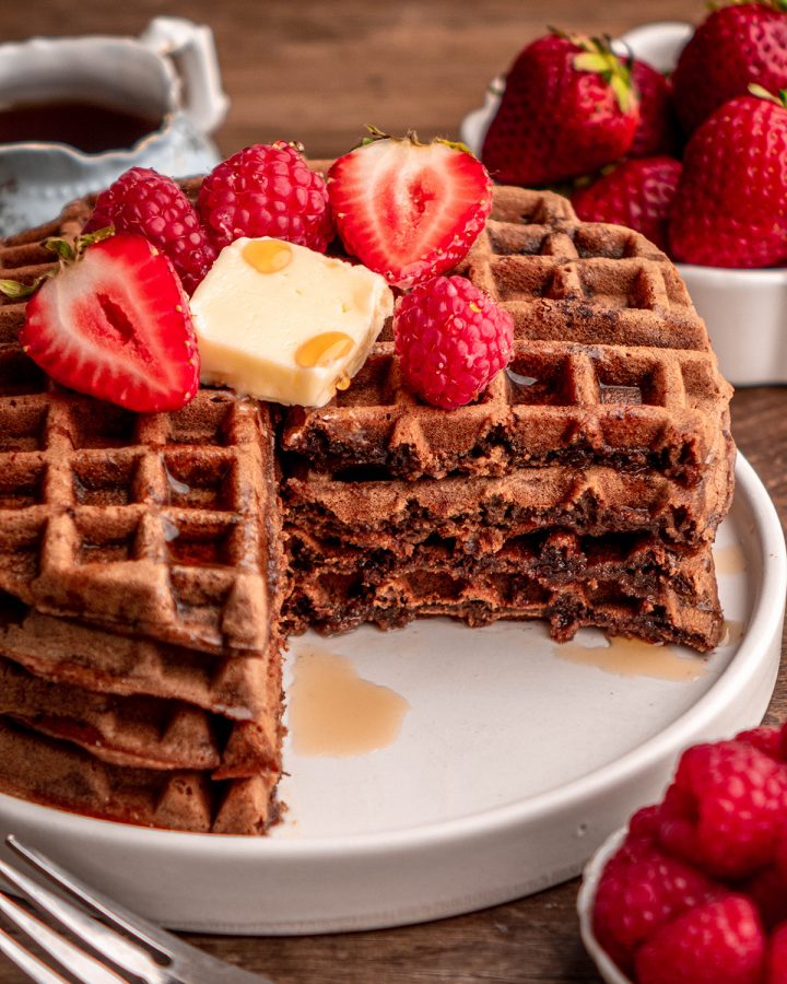a stack of 3 Chocolate Waffles with a piece cut out of it, butter, syrup and berries