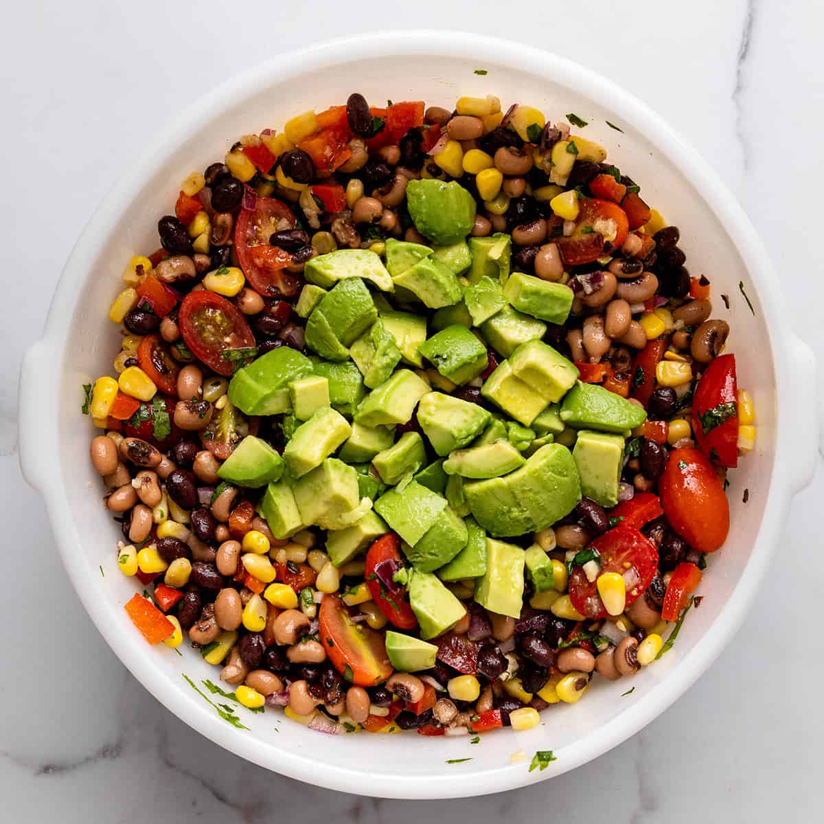 avocado being added to the cowboy caviar in a mixing bowl