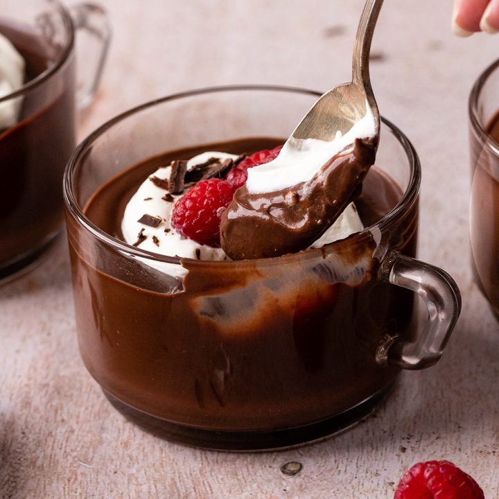 a spoon taking a scoop of Chocolate Pudding in a glass cup with whipped cream, raspberries, and chocolate shavings