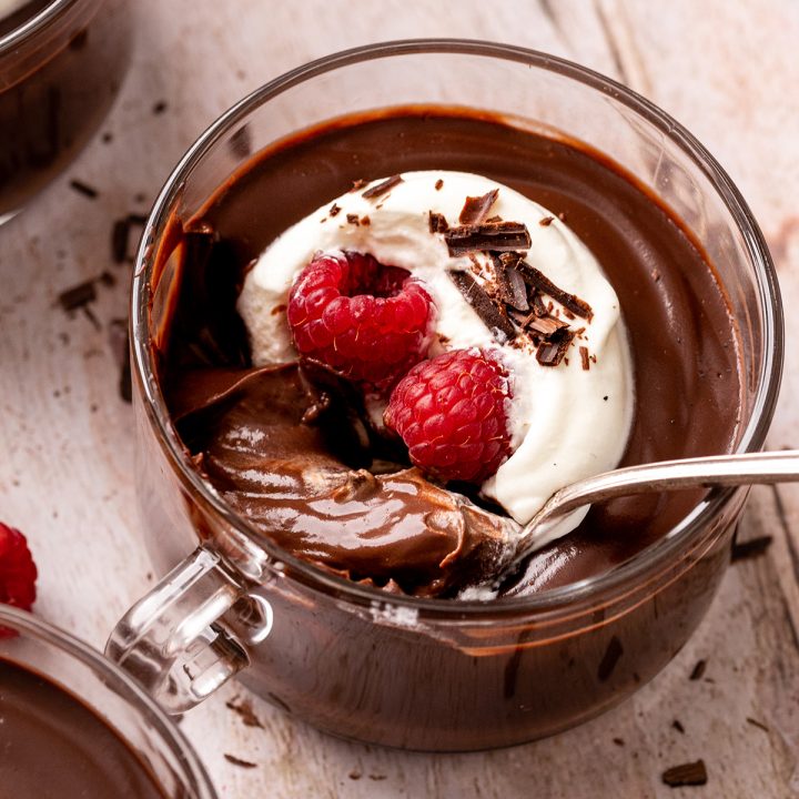 a spoon taking a scoop of Chocolate Pudding in a glass cup with whipped cream, raspberries, and chocolate shavings
