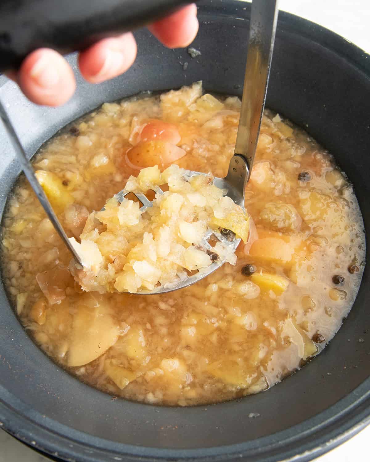how to make apple cider recipe - a hand holding a potato masher mashing the apples