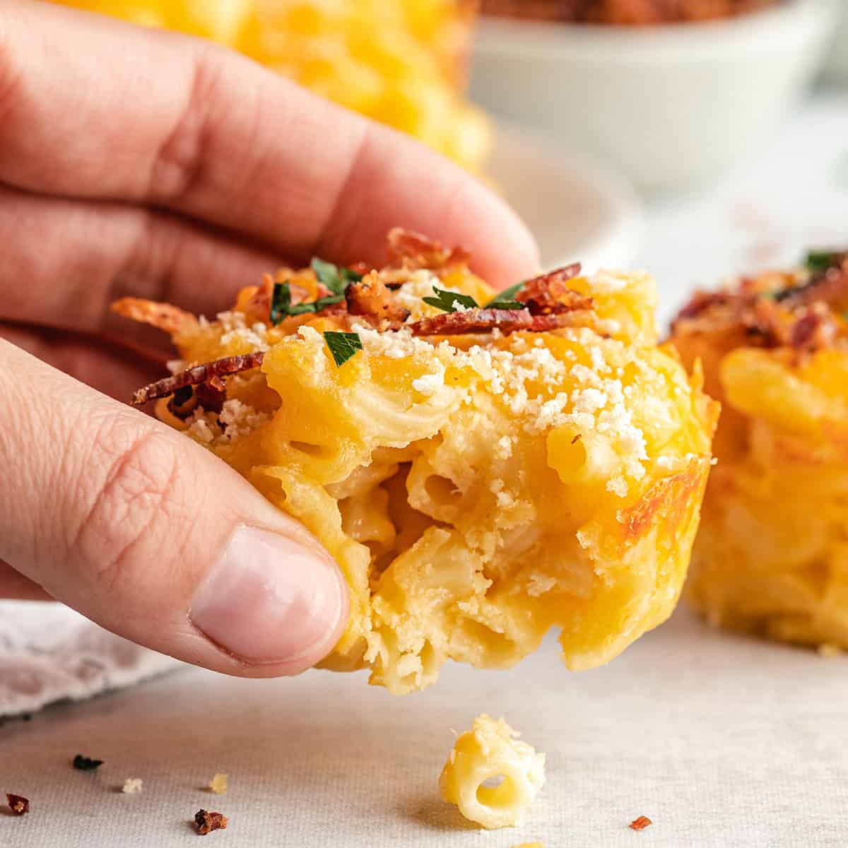a hand holding a baked macaroni and cheese cup with a bite taken out of it so the cheesy inside is visible. It's topped with cooked bacon, a breadcrumb topping and chopped fresh parsley. There are more mac and cheese cups in the background