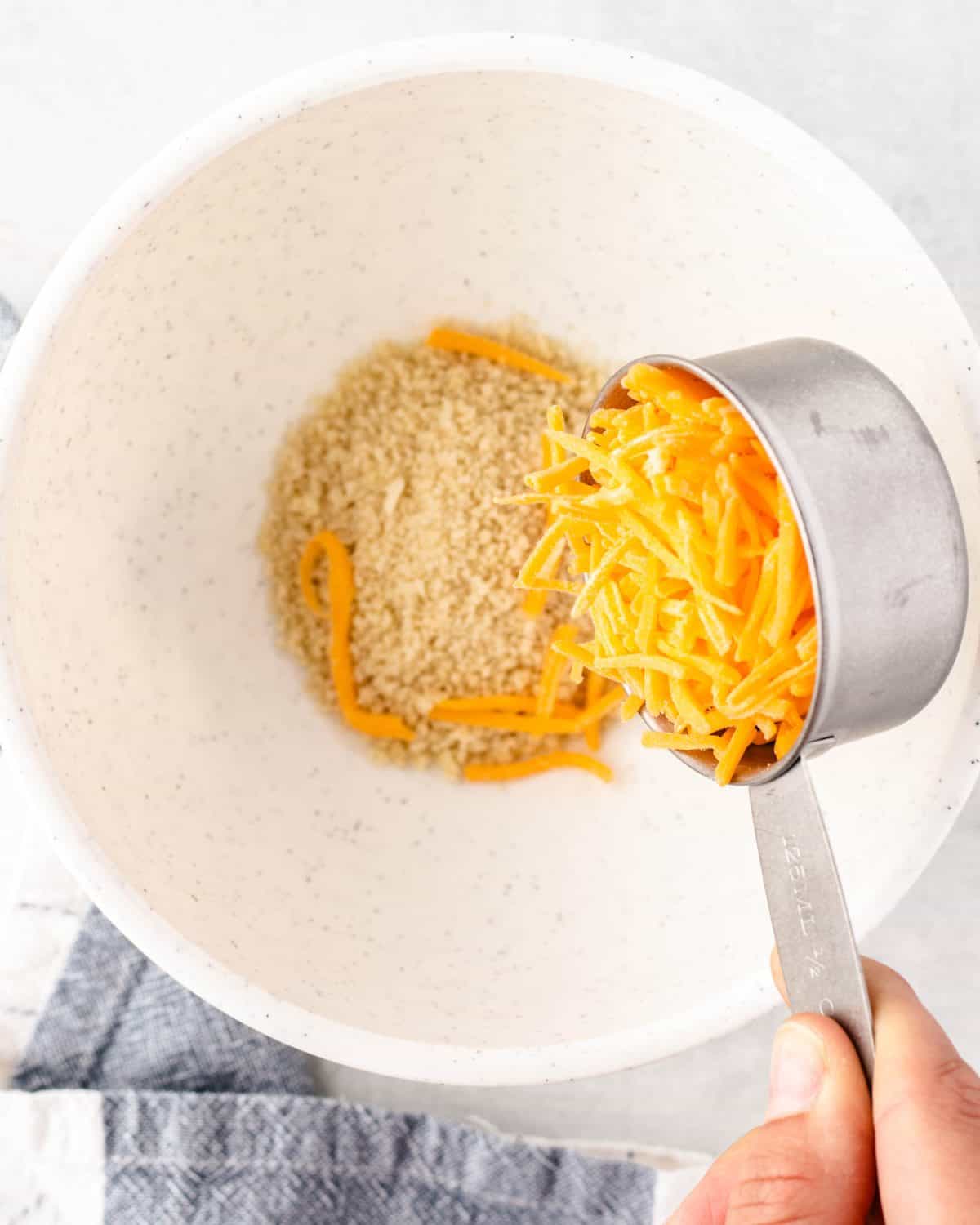 making mac and cheese cups topping - shredded cheddar cheese being poured into a white bowl with breadcrumbs in it