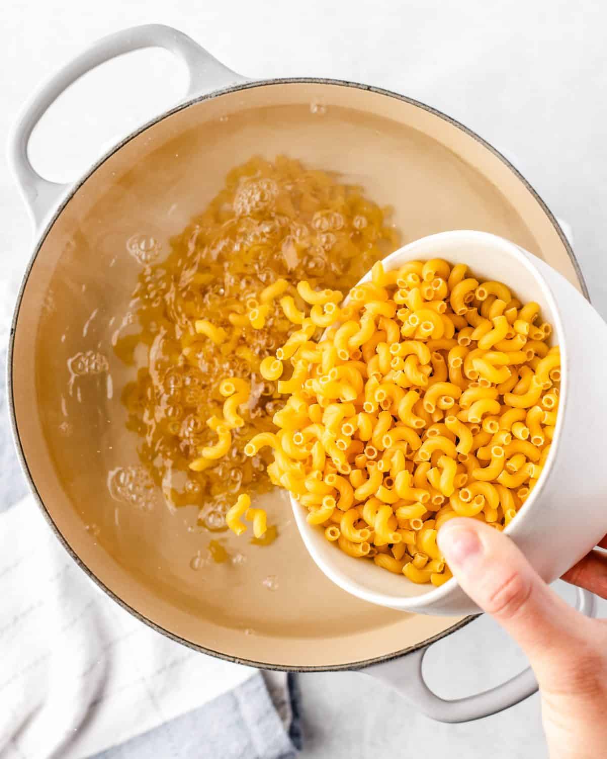 making mac and cheese cups - uncooked elbow macaroni noodles being poured from a white bowl into a white pot full of boiling water