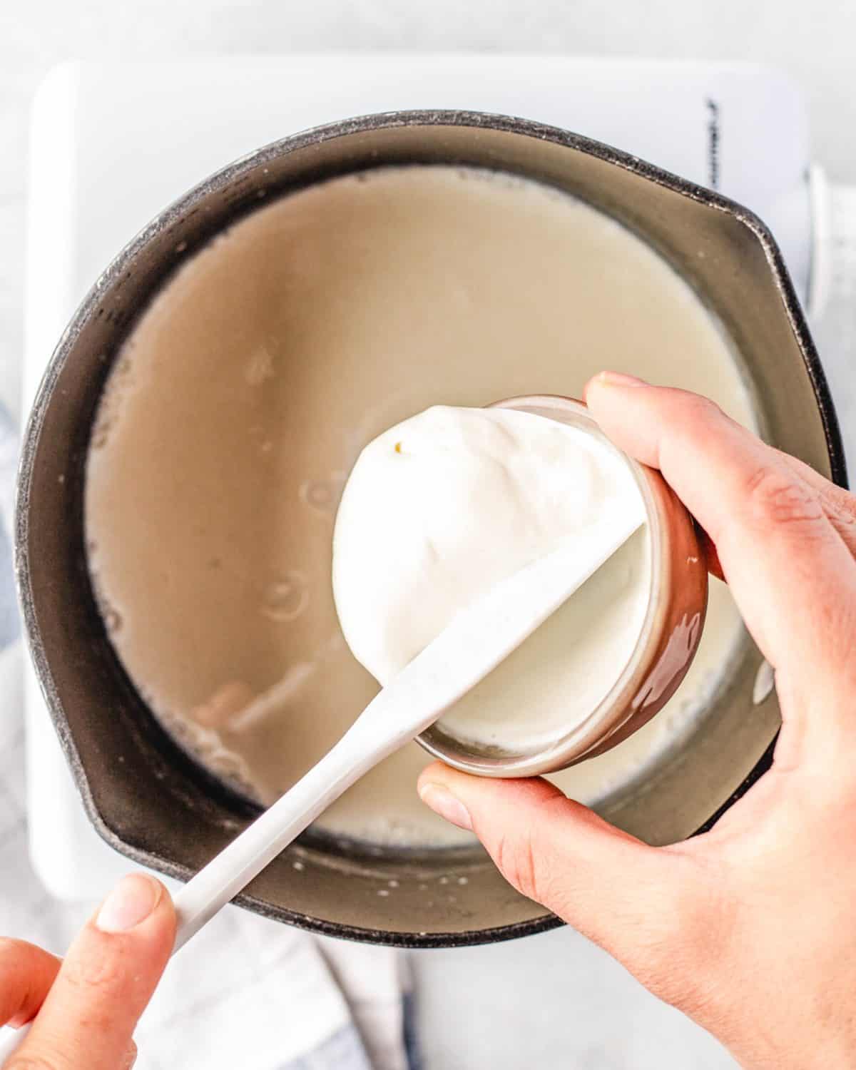 making macaroni and cheese cups sauce - sour cream being poured from a small bowl with a white spatula into the cast iron skillet