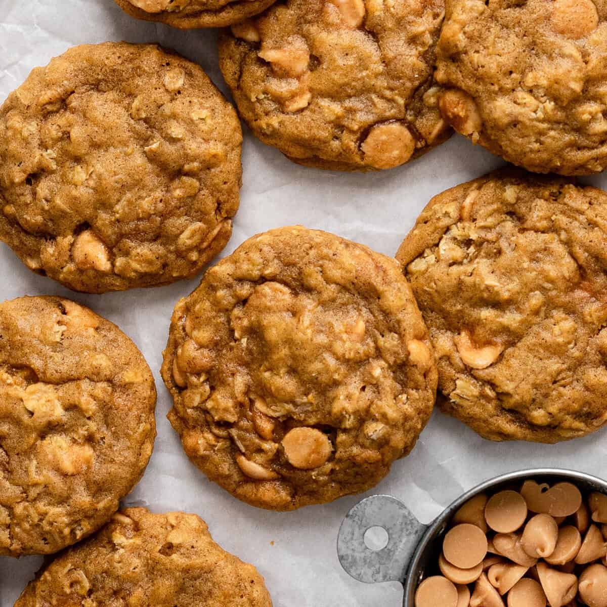 7 pumpkin oatmeal cookies on parchment paper with butterscotch chips in a silver measuring cup nearby