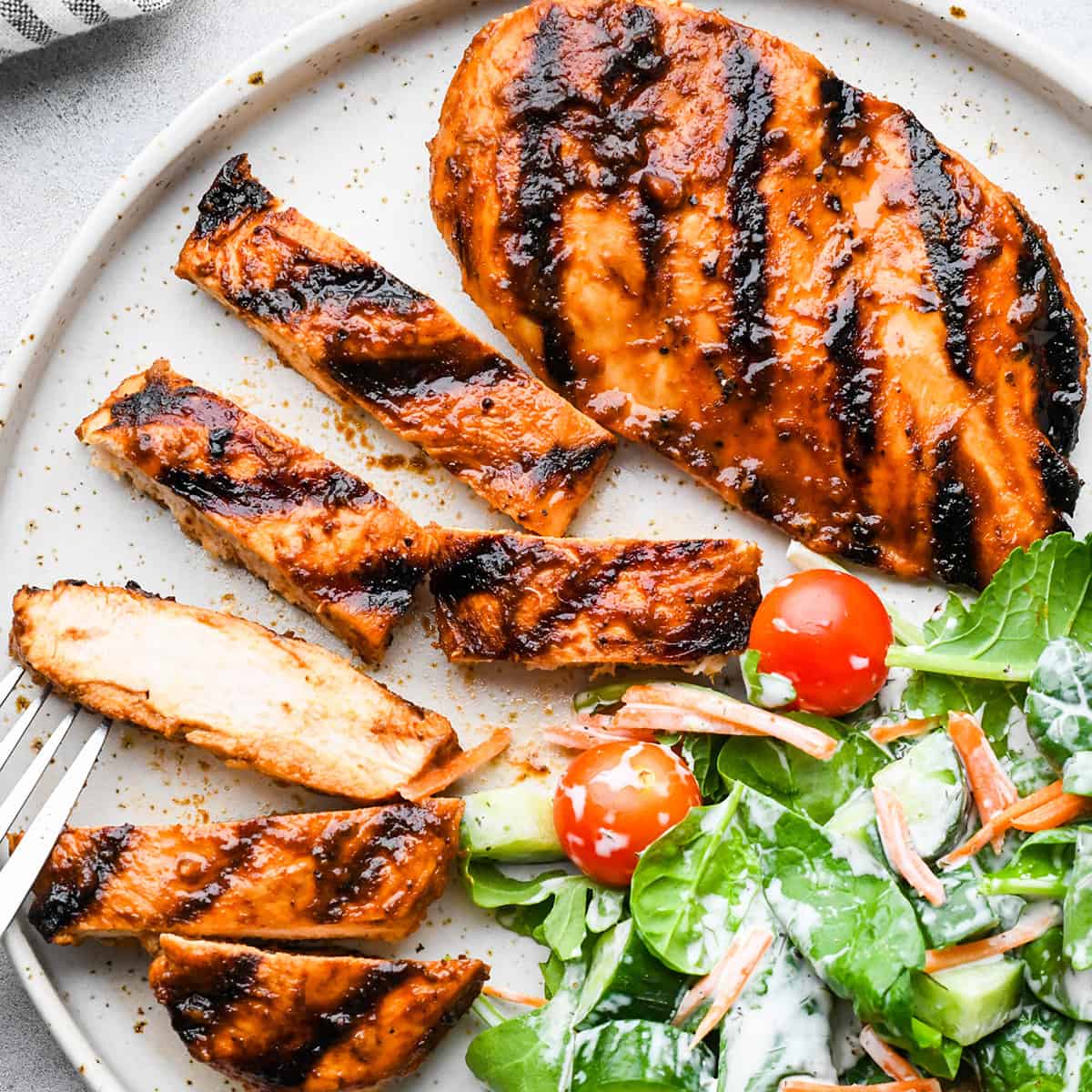 2 grilled bbq chicken breasts on a plate with a salad, one cut into 6 strips