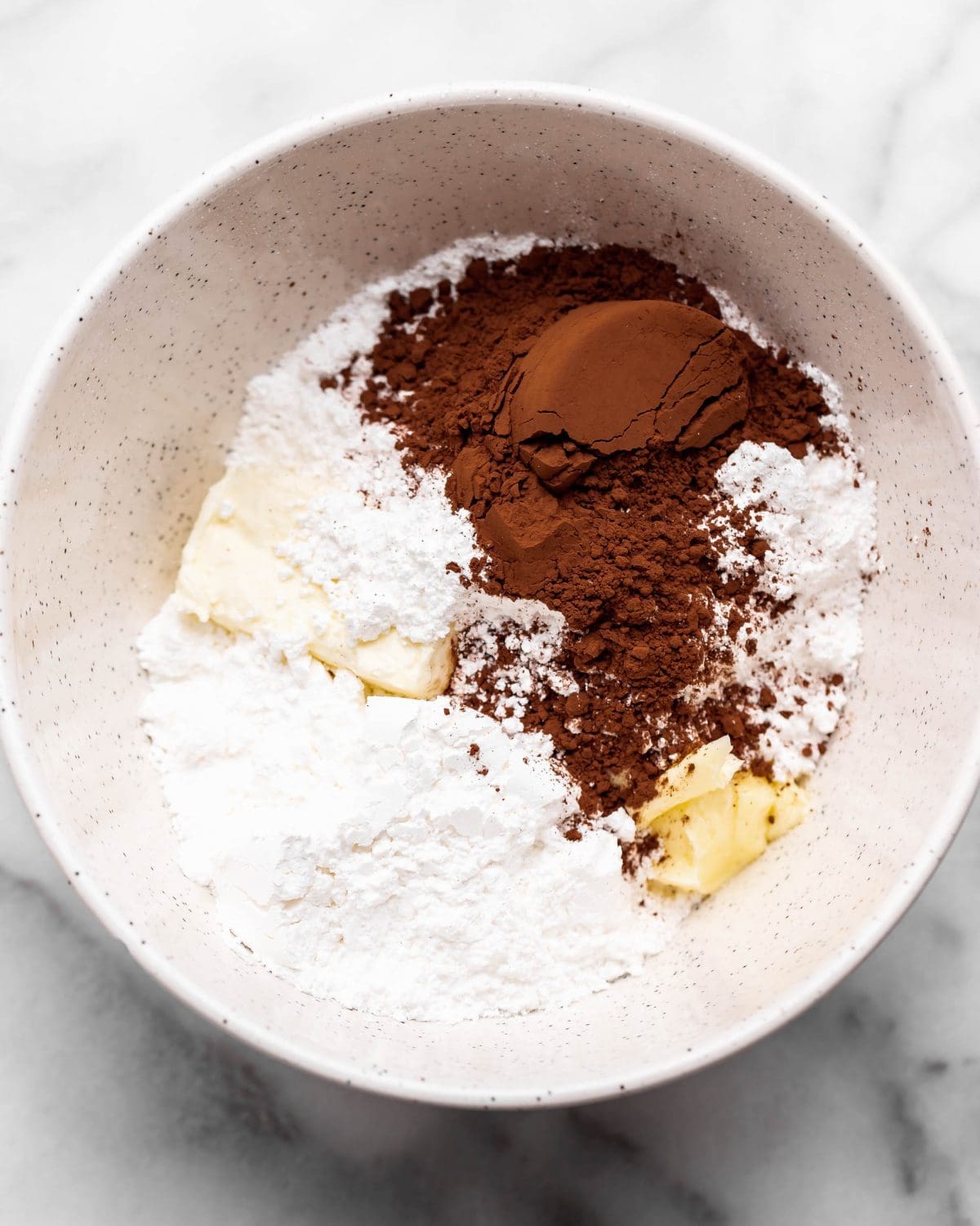 making cream cheese frosting for chocolate cinnamon rolls - butter, powdered sugar, cream cheese and cocoa powder in a large white ceramic mixing bowl before mixing