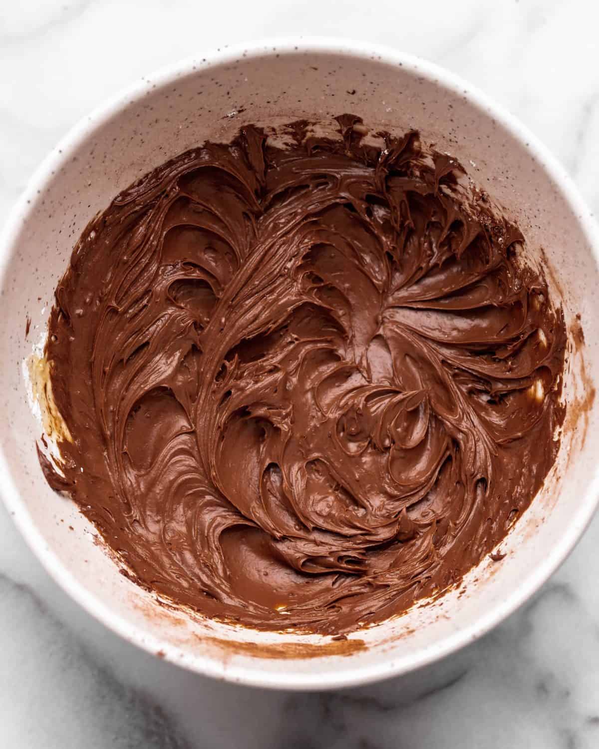 chocolate cream cheese frosting for chocolate cinnamon rolls in a large white ceramic mixing bowl on a marble surface