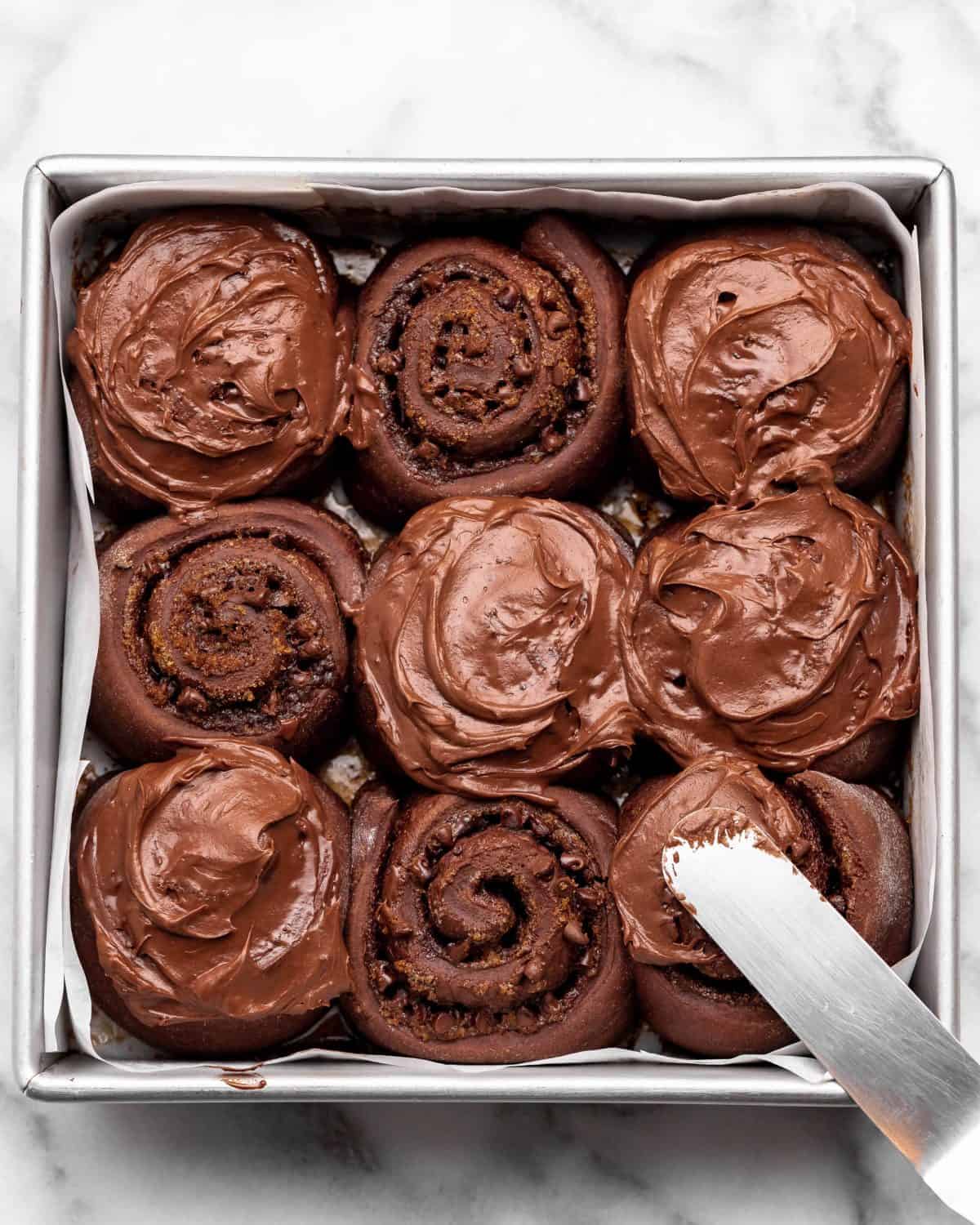 9 chocolate cinnamon rolls in a square metal baking dish lined with parchment paper after baking 6 chocolate cinnamon rolls have chocolate frosting on them, the one in the bottom right corner is being frosting with a metal spatula
