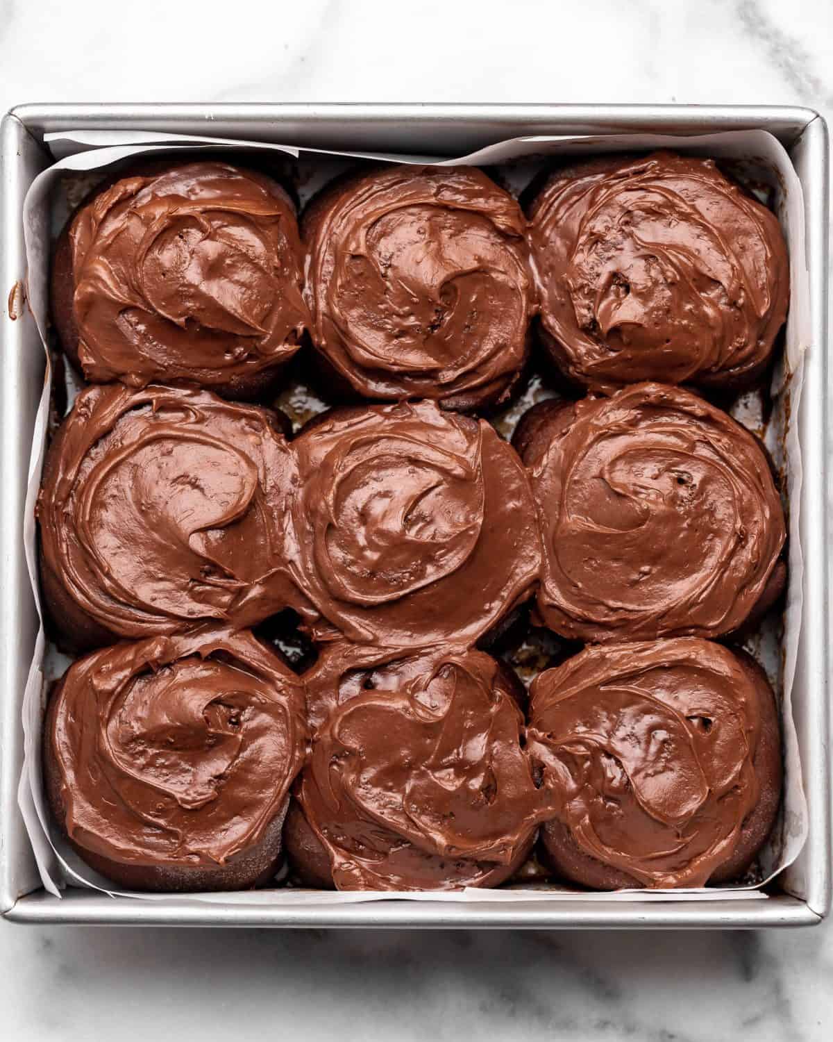 9 chocolate cinnamon rolls with chocolate frosting in a square metal baking dish lined with parchment paper.