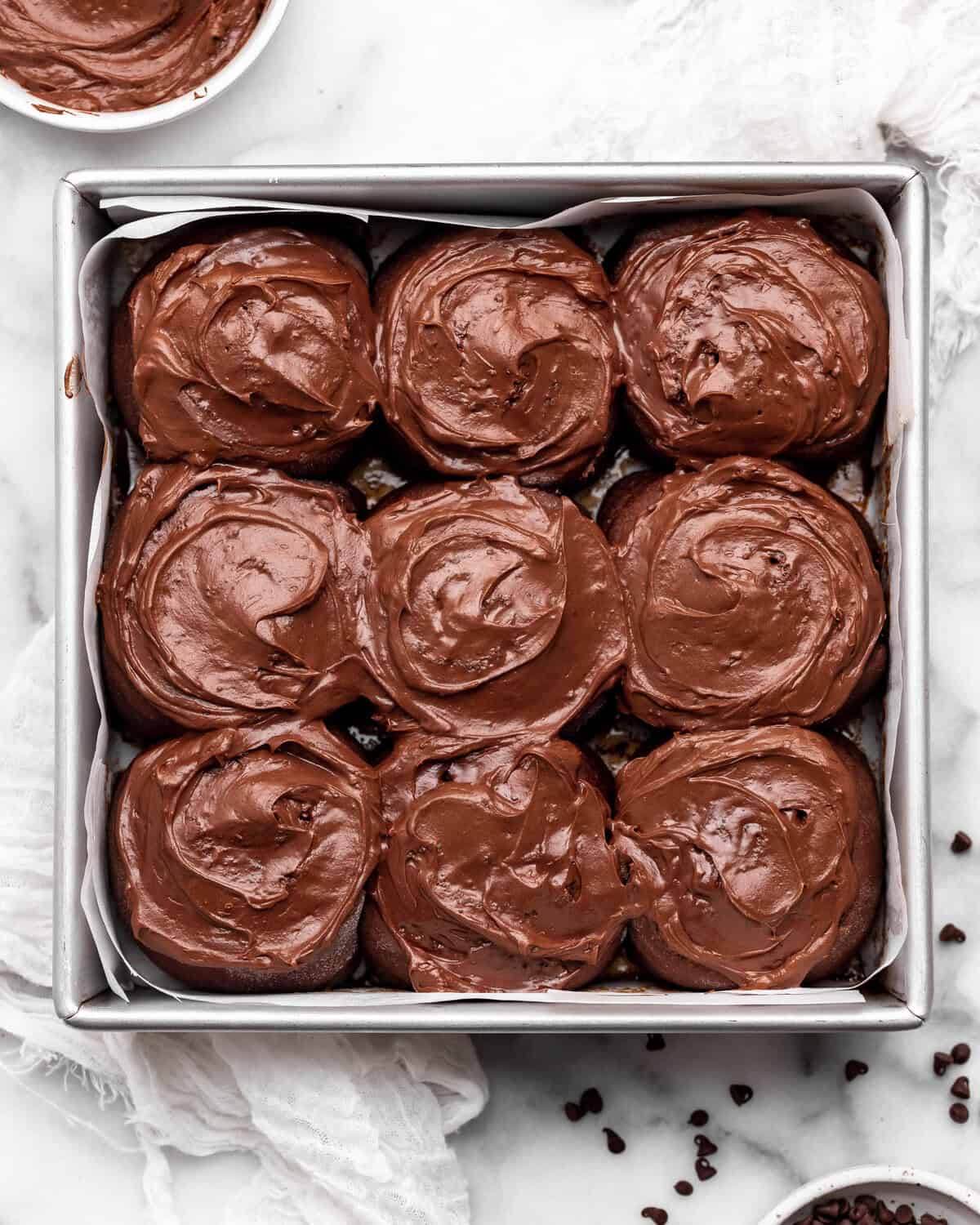 9 chocolate cinnamon rolls with chocolate frosting in a square metal baking dish lined with parchment paper. there is a bowl of chocolate frosting in the top left corner and mini chocolate chips around the pan.