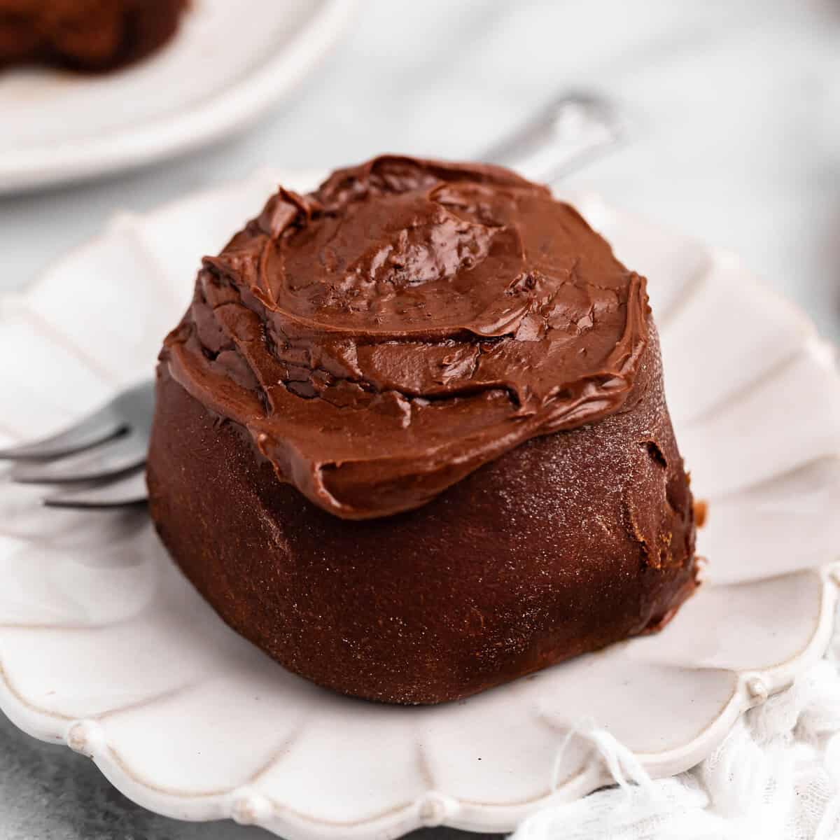 one Chocolate Cinnamon Roll on a fluted white ceramic plate topped with chocolate frosting. There's a silver fork on the plate and another plate with a cinnamon roll on it in the background
