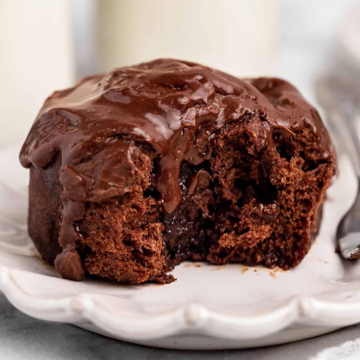one chocolate cinnamon rolls with chocolate frosting on top, on a white scalloped plate with a silver fork. There is a bite taken out of the cinnamon roll so the gooey texture is visible.