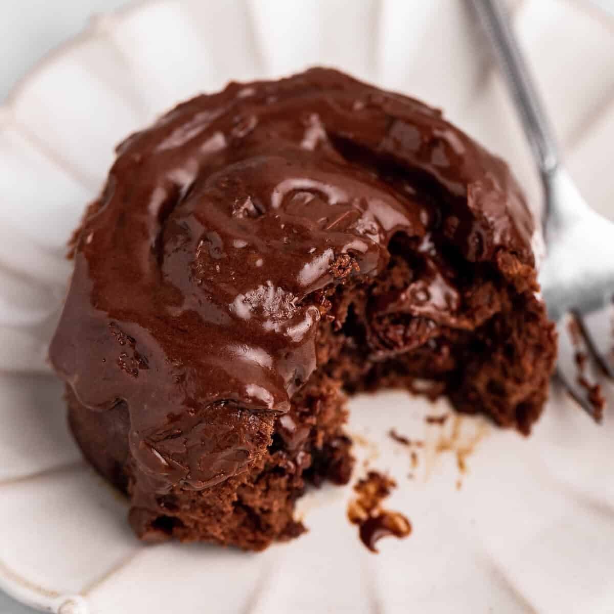 one chocolate cinnamon rolls with chocolate frosting on top, on a white scalloped plate with a silver fork. There is a bite taken out of the cinnamon roll so the gooey texture is visible.