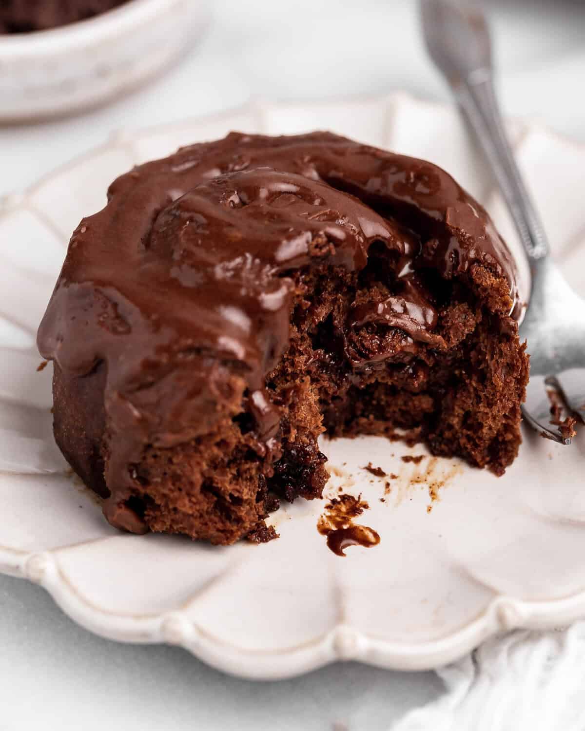 one chocolate cinnamon rolls with chocolate frosting on top, on a white scalloped plate with a silver fork. There is a bite taken out of the cinnamon roll so the gooey texture is visible.