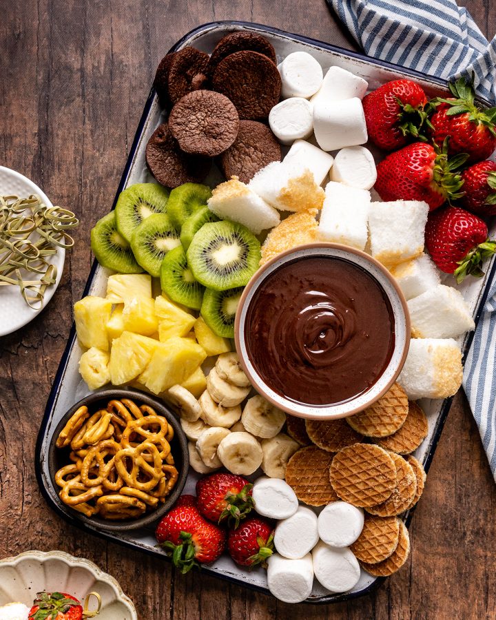 a bowl of Chocolate Fondue surrounded by fruit, cake, brownies, cookies and pretzels