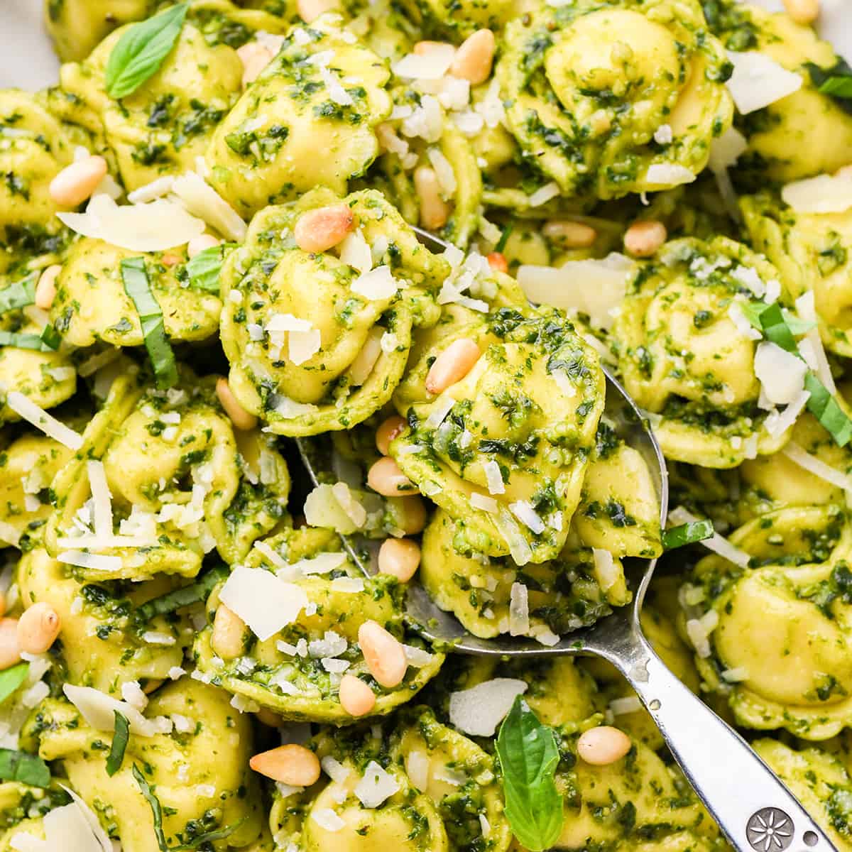a spoon scooping up pesto tortellini topped with parmesan and pine nuts 