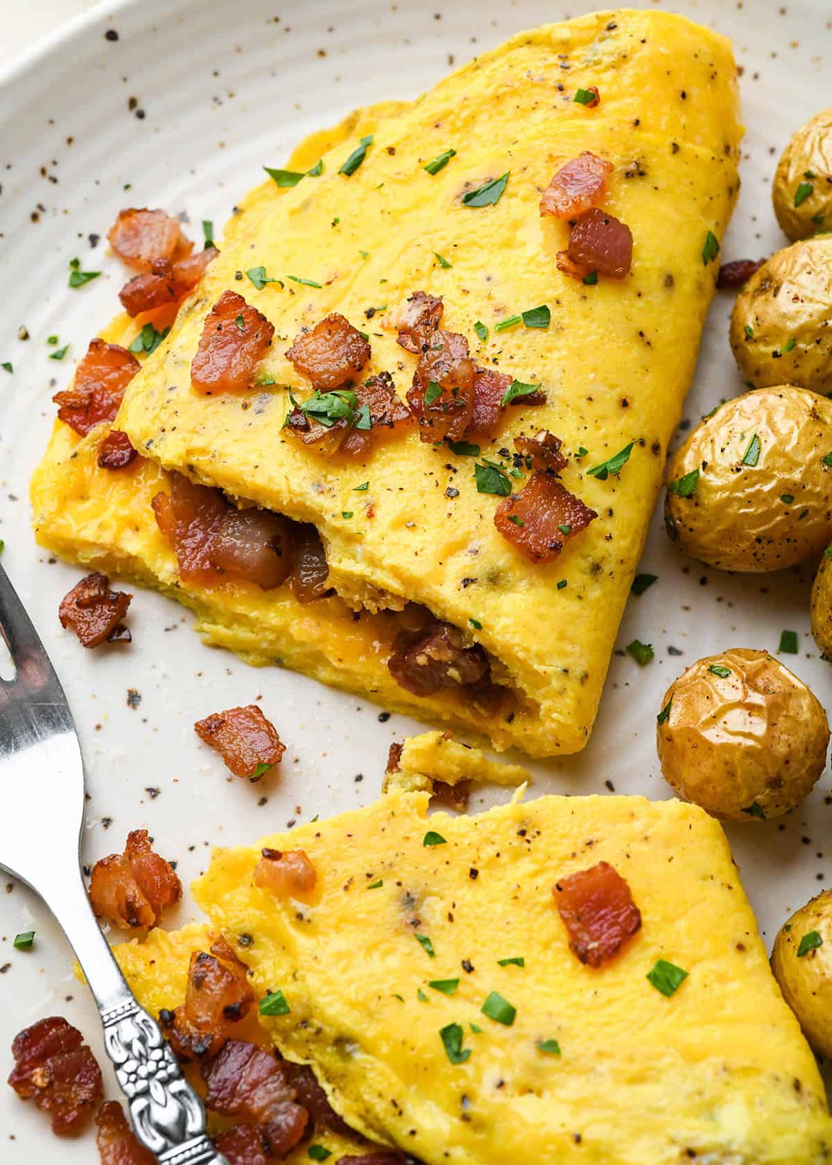 a fluffy omelette on a white ceramic plate with a bite taken out of it with a silver fork, so you can see that it's filled with cheese and bacon and topped with bacon and chopped parsley, there are roasted potatoes on the plate as well.