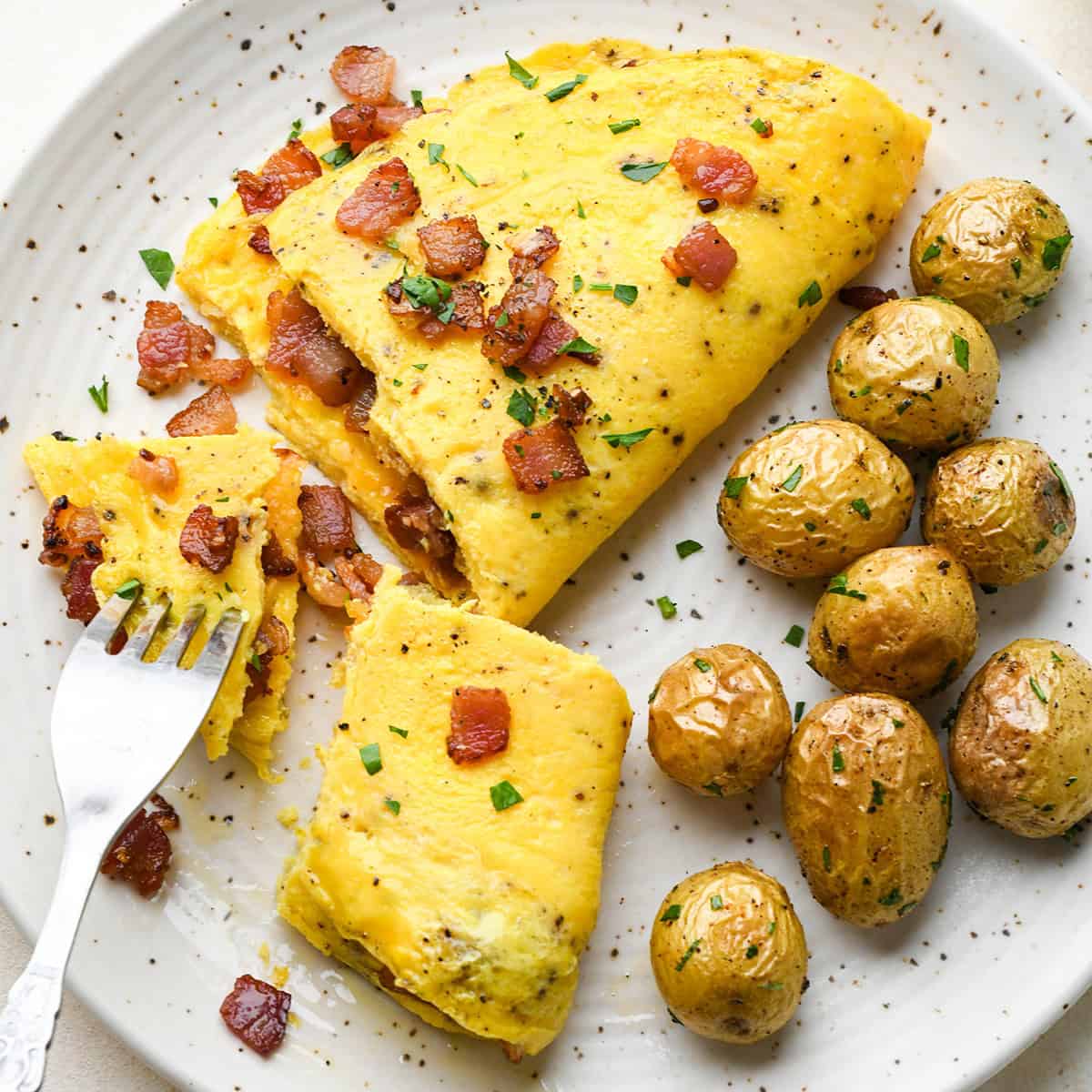 a fluffy omelette on a white ceramic plate with two bites taken out of it with a silver fork, so you can see that it's filled with cheese and bacon and topped with bacon and chopped parsley, there are roasted potatoes on the plate as well.