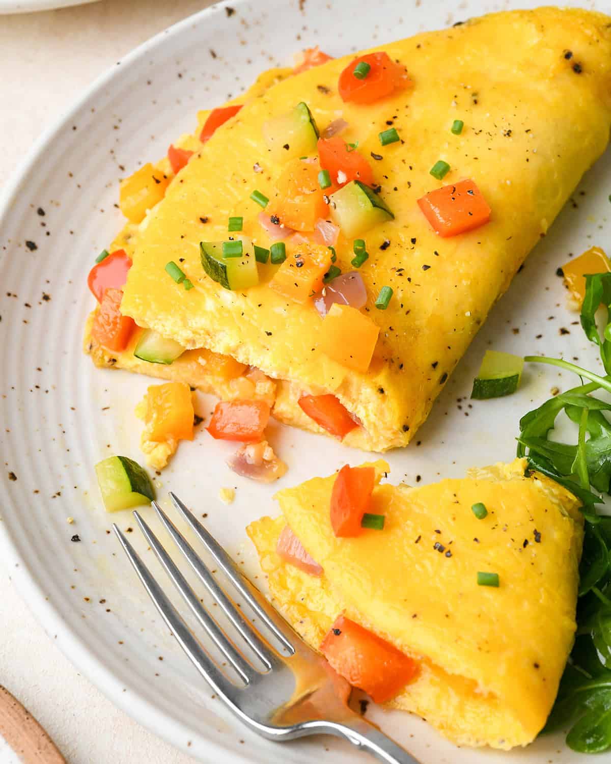 a fluffy omelette recipe on a white ceramic plate filled with vegetables (bell peppers, zucchini and onions) and cheese, and topped with cooked veggies and sliced chives. A fork is taking a bite out of it, and there is arugula on the plate in the bottom right corner.