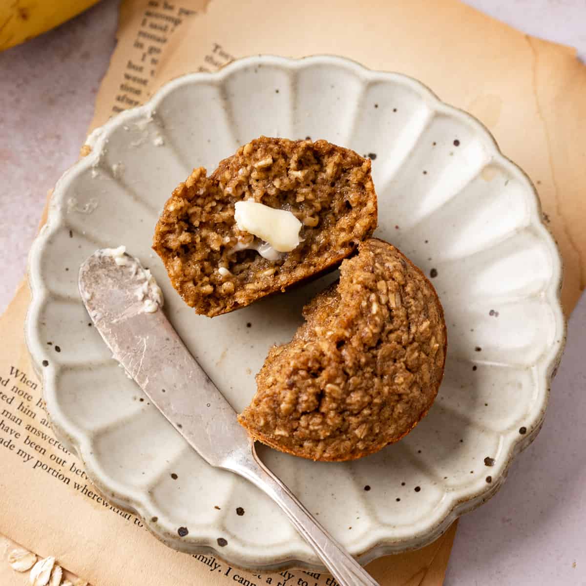 a Banana Oatmeal Muffin on a plate cut in half with butter on one half. 
