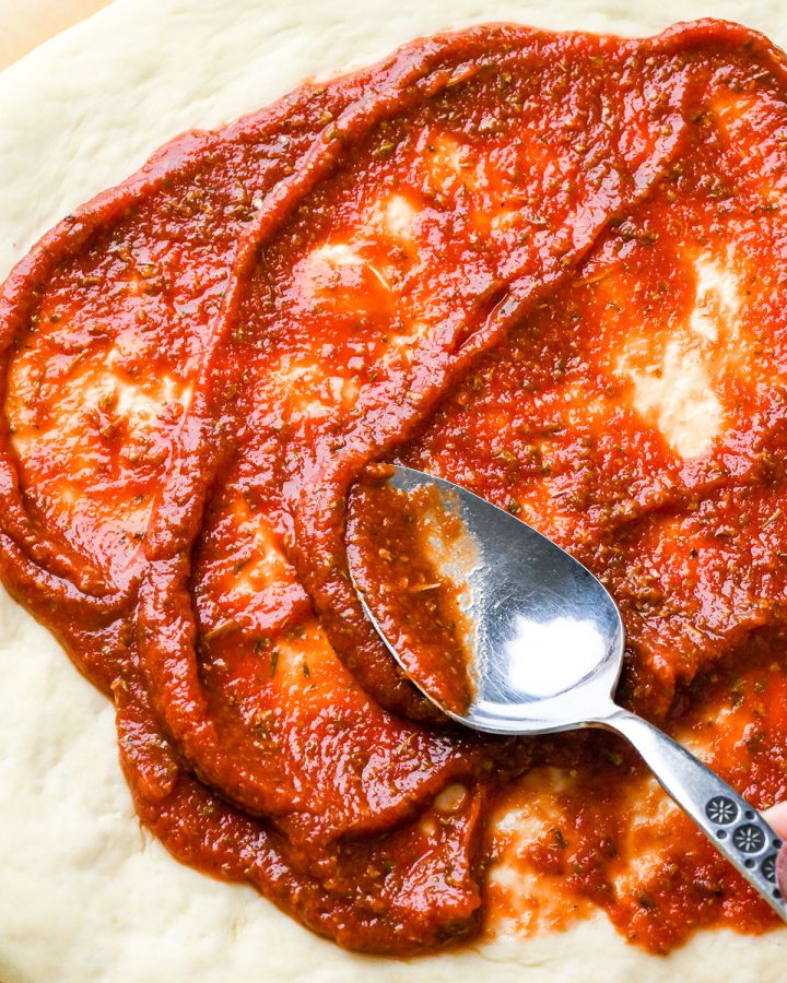 up close photo of pizza sauce being spread onto pizza dough with a spoon