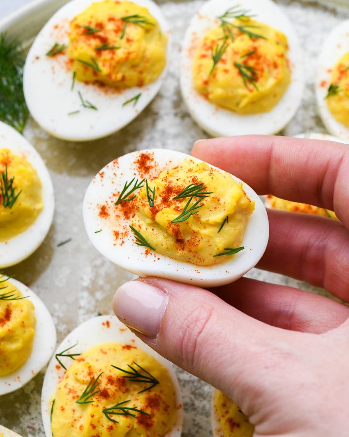 a hand holding a Deviled Egg garnished with dill and paprika