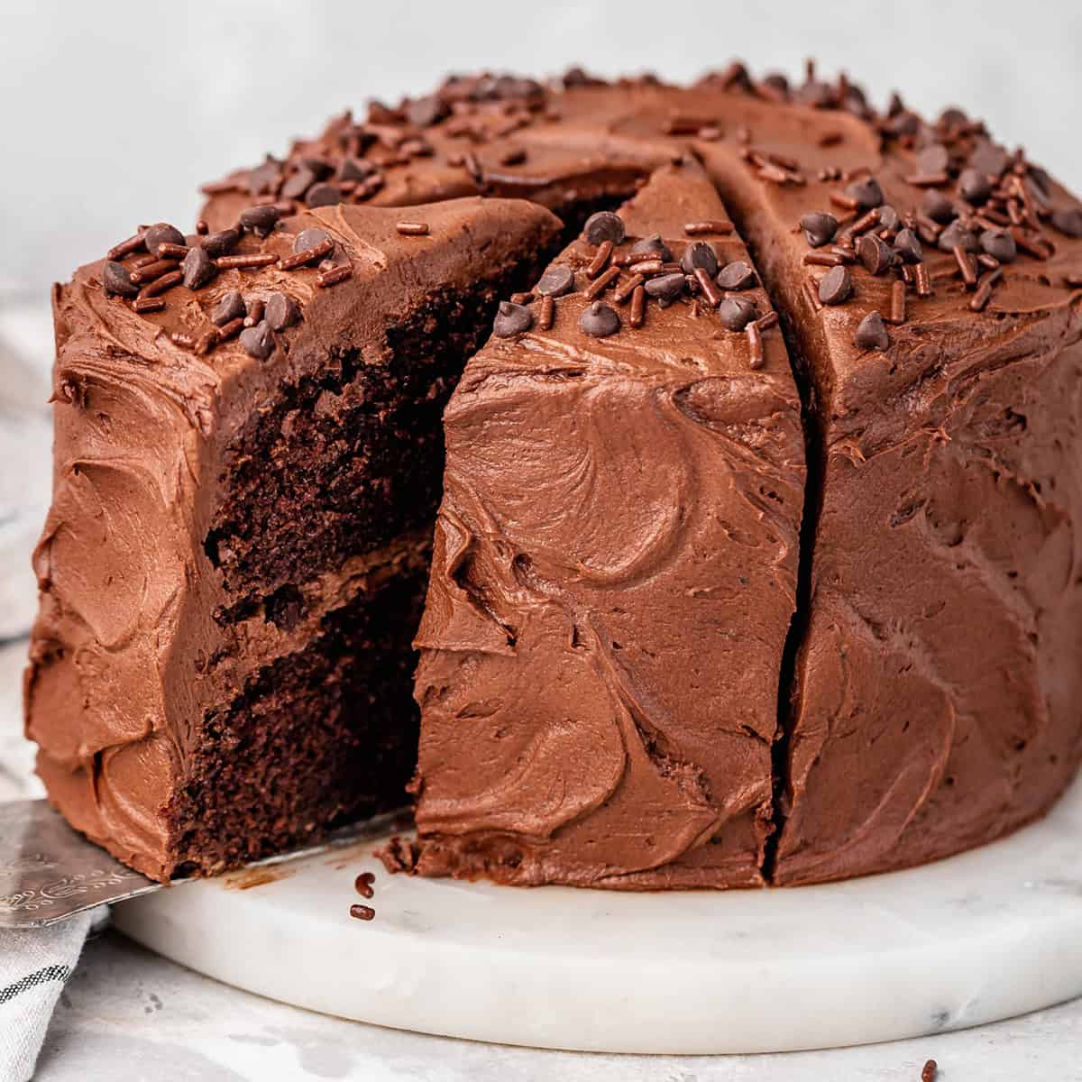 a whole frosted Chocolate Cake with chocolate frosting on a white marble cake plate topped with chocolate chips and sprinkles two slices have been cut out, and one is being pulled off the plate with a metal spatula