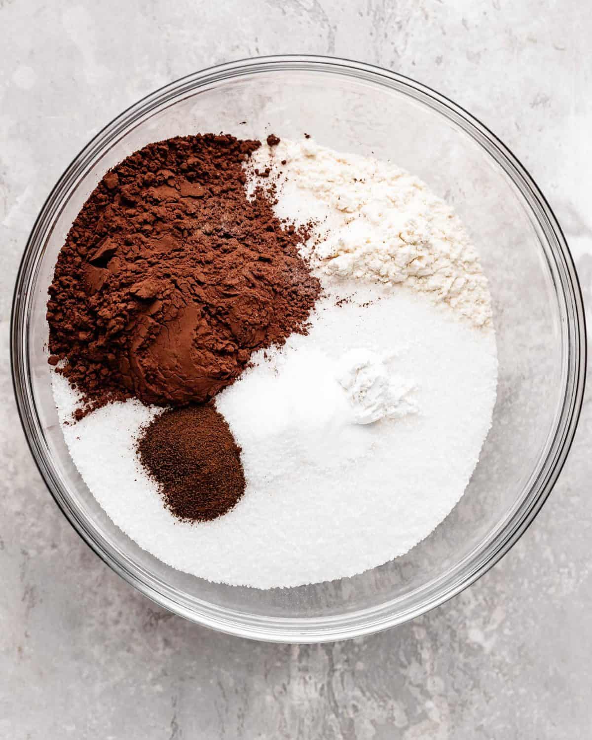 how to make chocolate cake recipe - flour, sugar, cocoa powder, baking soda, baking powder, sea salt and espresso powder in a glass bowl on a gray surface before mixing