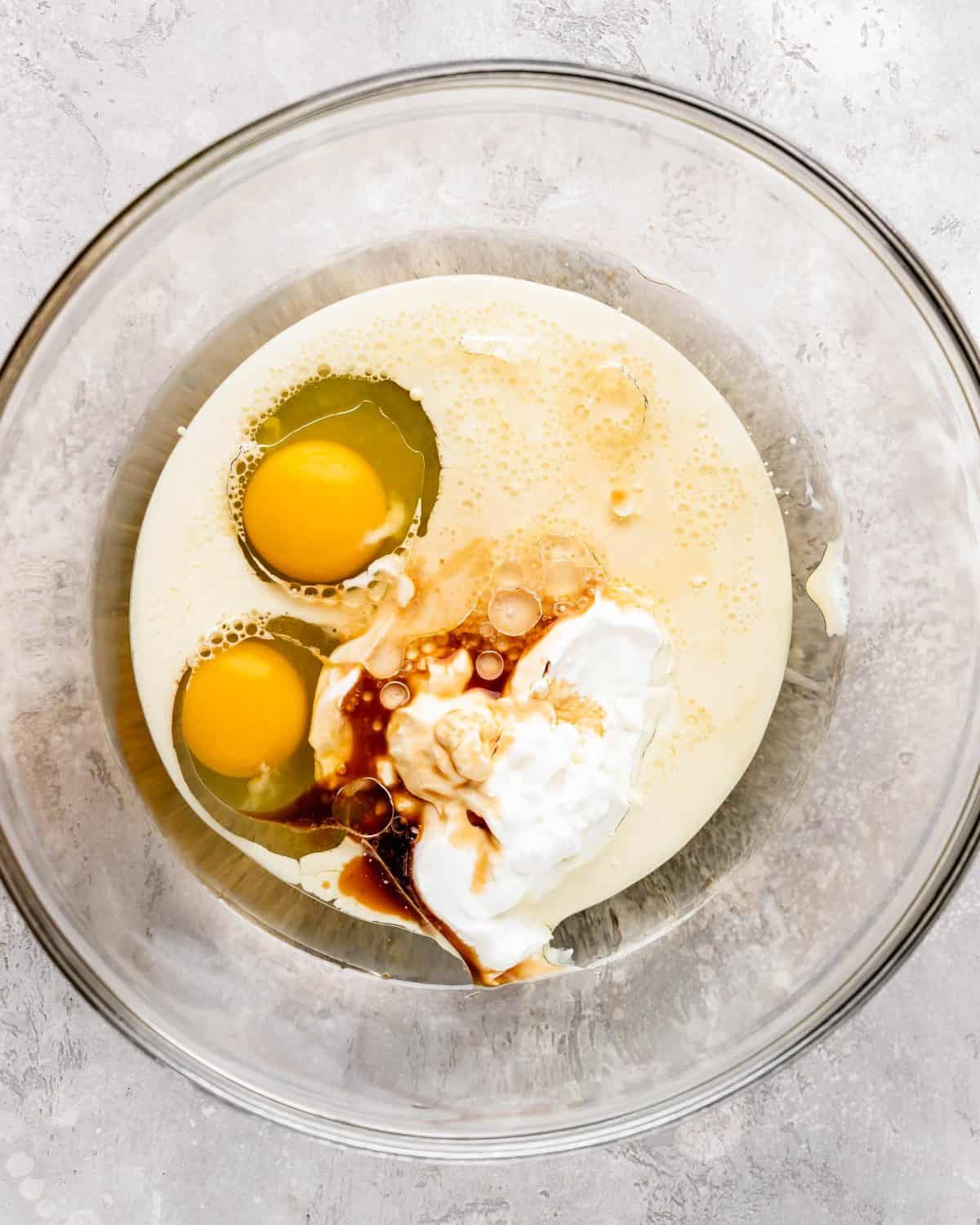 how to make chocolate cake from scratch - eggs, milk, canola oil, sour cream and vanilla in a large glass bowl on a gray surface before mixing