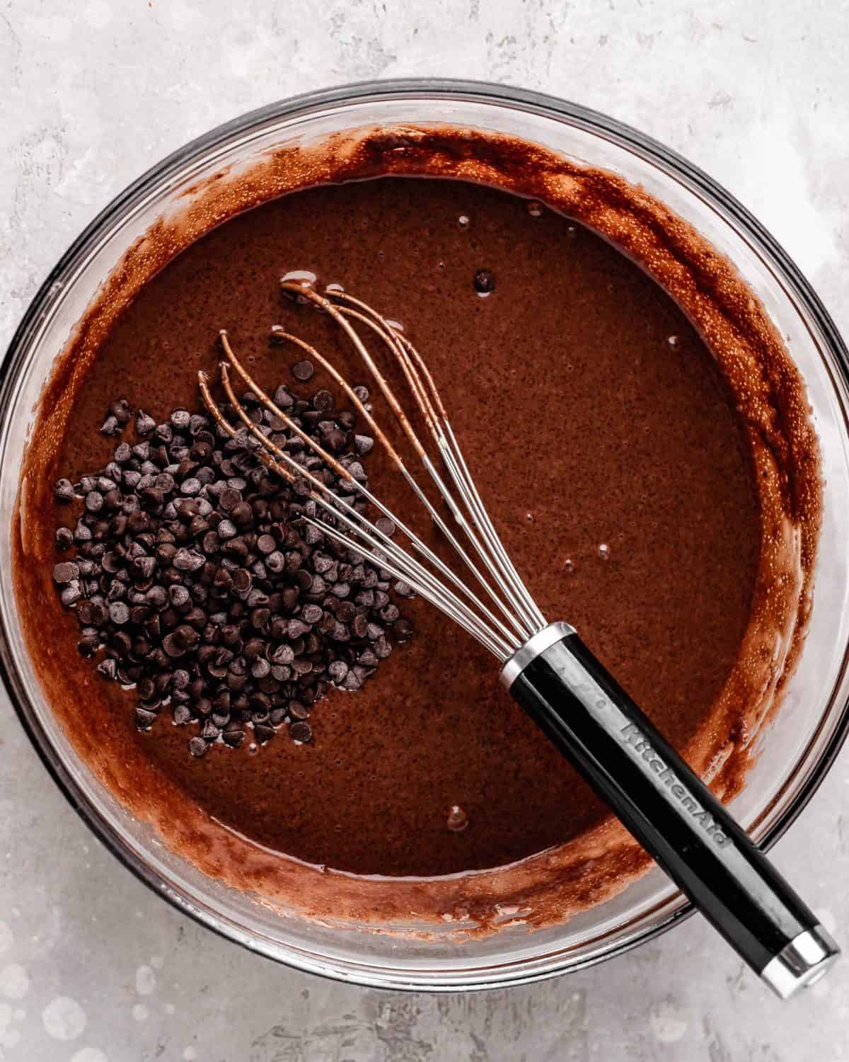 how to make chocolate cake from scratch - mini chocolate chips added to the chocolate cake batter in a large glass bowl with a metal whisk before stirring
