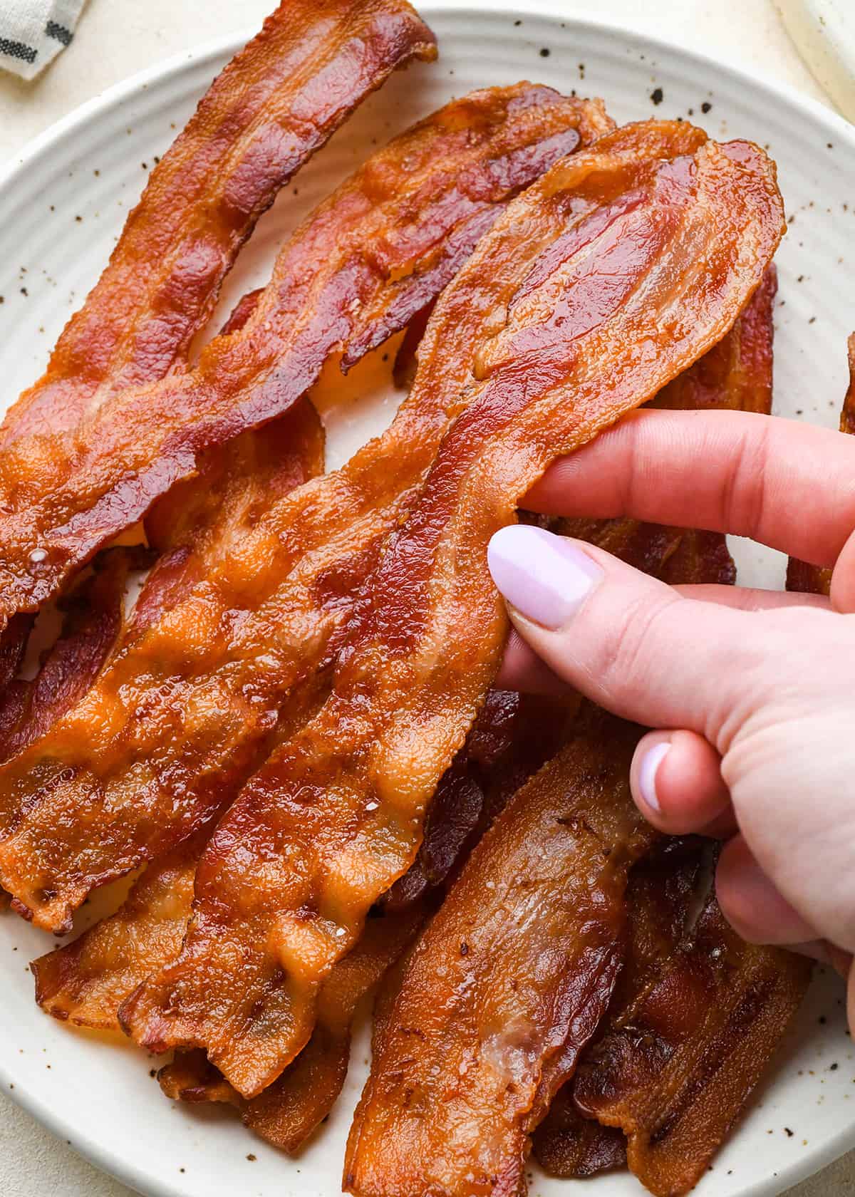 a hand picking up a piece of Oven Baked Bacon from a plate