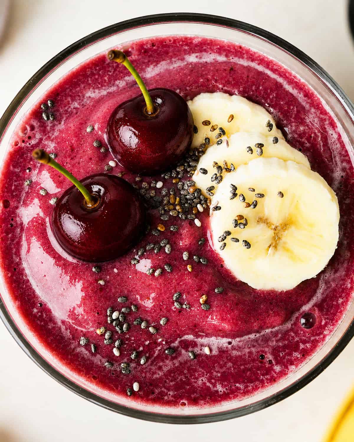 close up overhead photo of cherry smoothie recipe in a glass topped with two cherries, 3 slices of banana and a sprinkle of chia seeds.