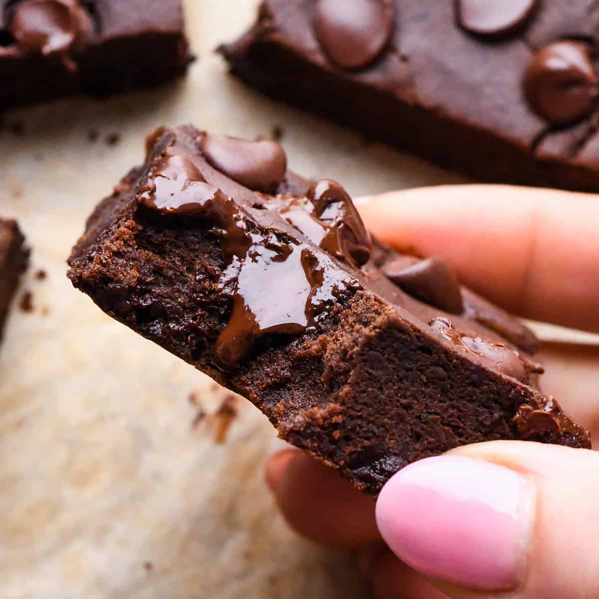 a hand holding a black bean brownie with a bite taken out of it so they fudgy texture is visible. There is melty chocolate chips dripping down the front of the brownie. It's being held over more brownies on parchment paper.