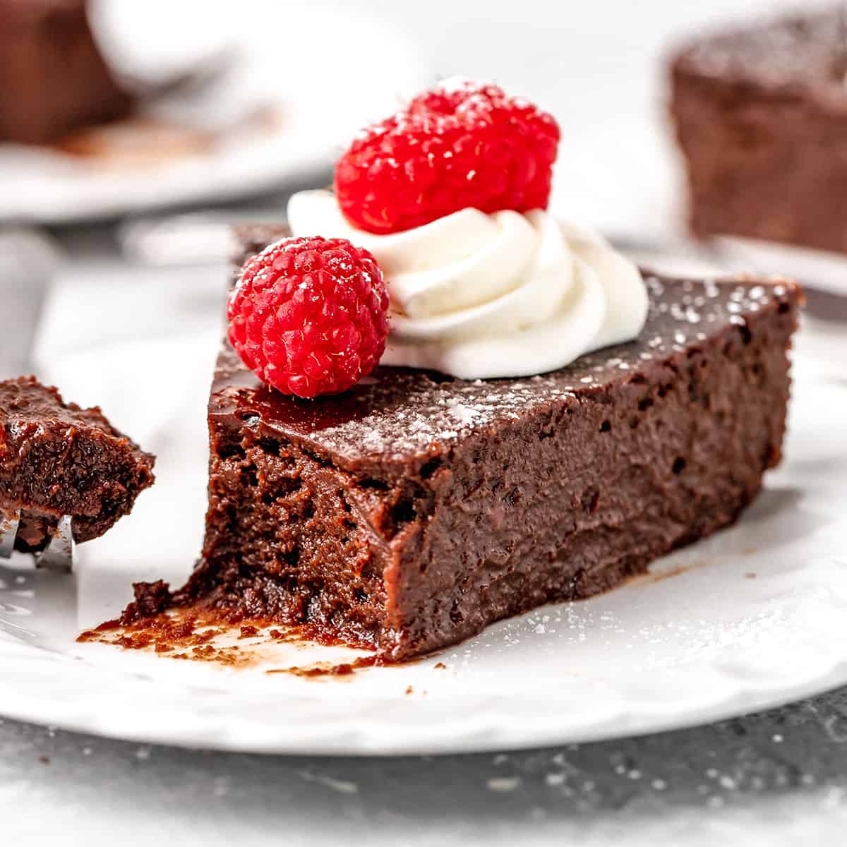 a piece of Flourless Chocolate Cake on a plate with a bite taken out of it, garnished with raspberries and whipped cream