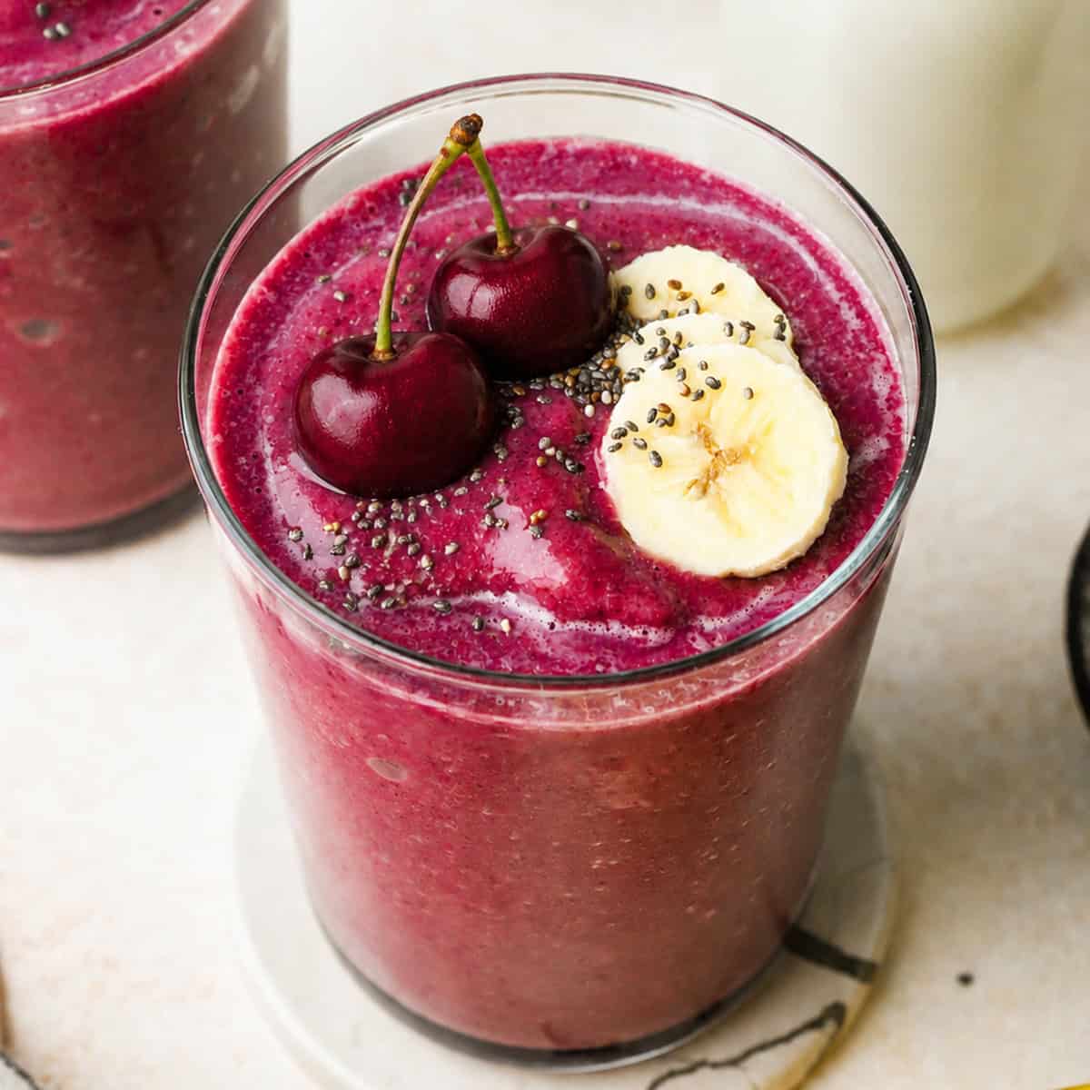 cherry smoothie recipe in a glass topped with two cherries, 3 slices of banana and a sprinkle of chia seeds. There is another glass of cherry fruit smoothie and a glass of milk in the background