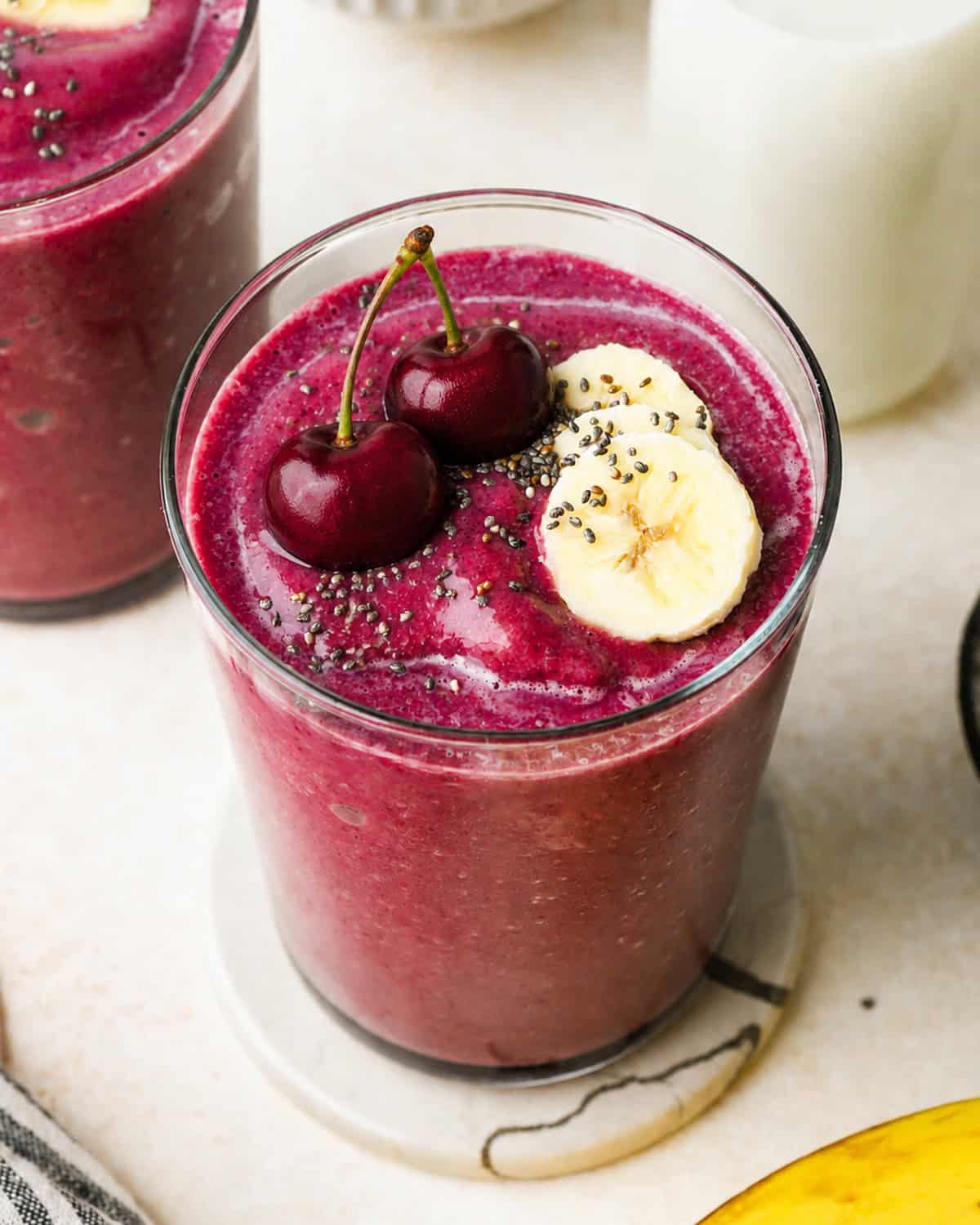 cherry smoothie recipe in a glass topped with two cherries, 3 slices of banana and a sprinkle of chia seeds. There is another glass of cherry fruit smoothie and a glass of milk in the background