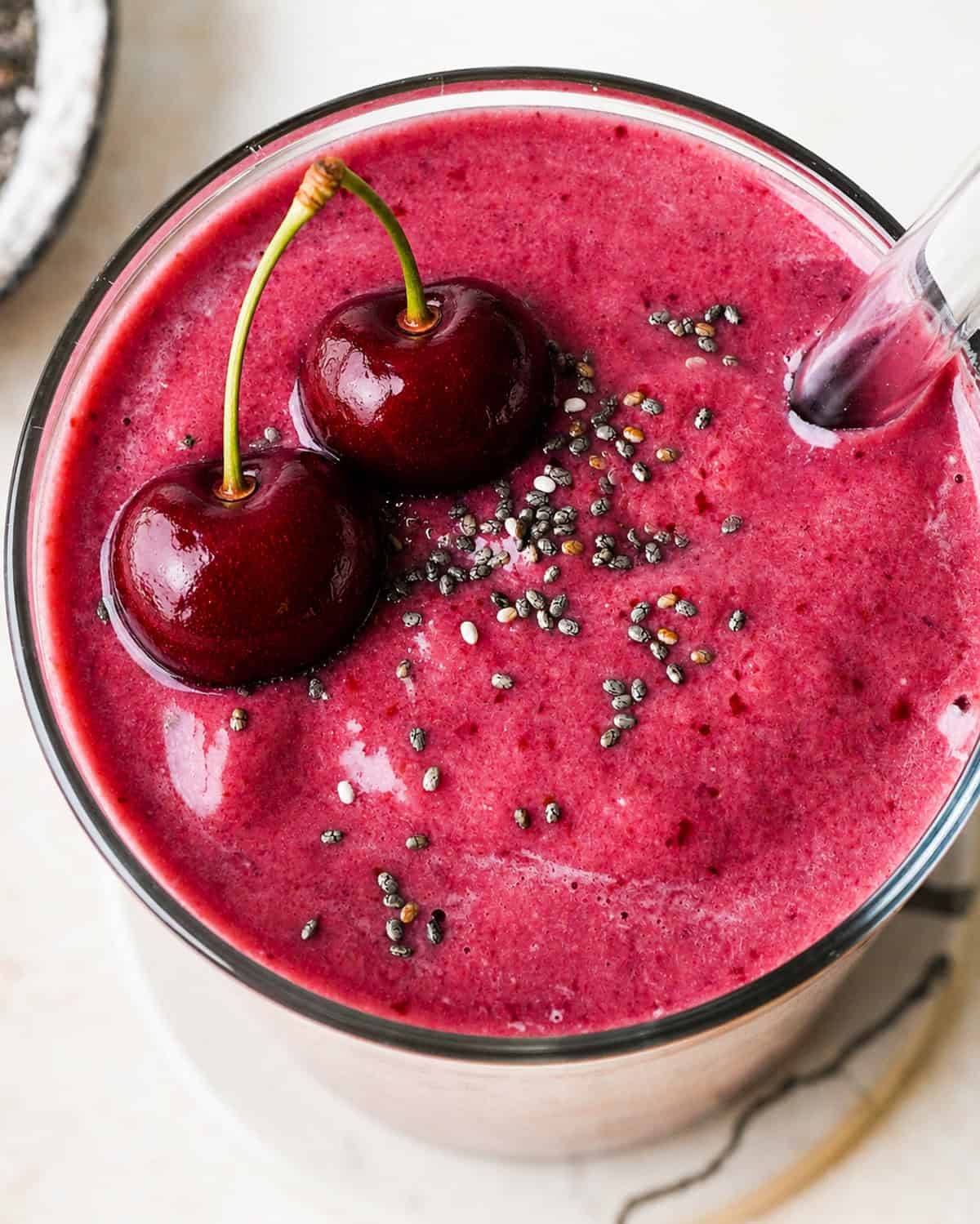 cherry smoothie recipe in a glass topped with two cherries, and a sprinkle of chia seeds. There is a glass straw in the smoothie. There is a bowl of chia seeds in the top left corner.