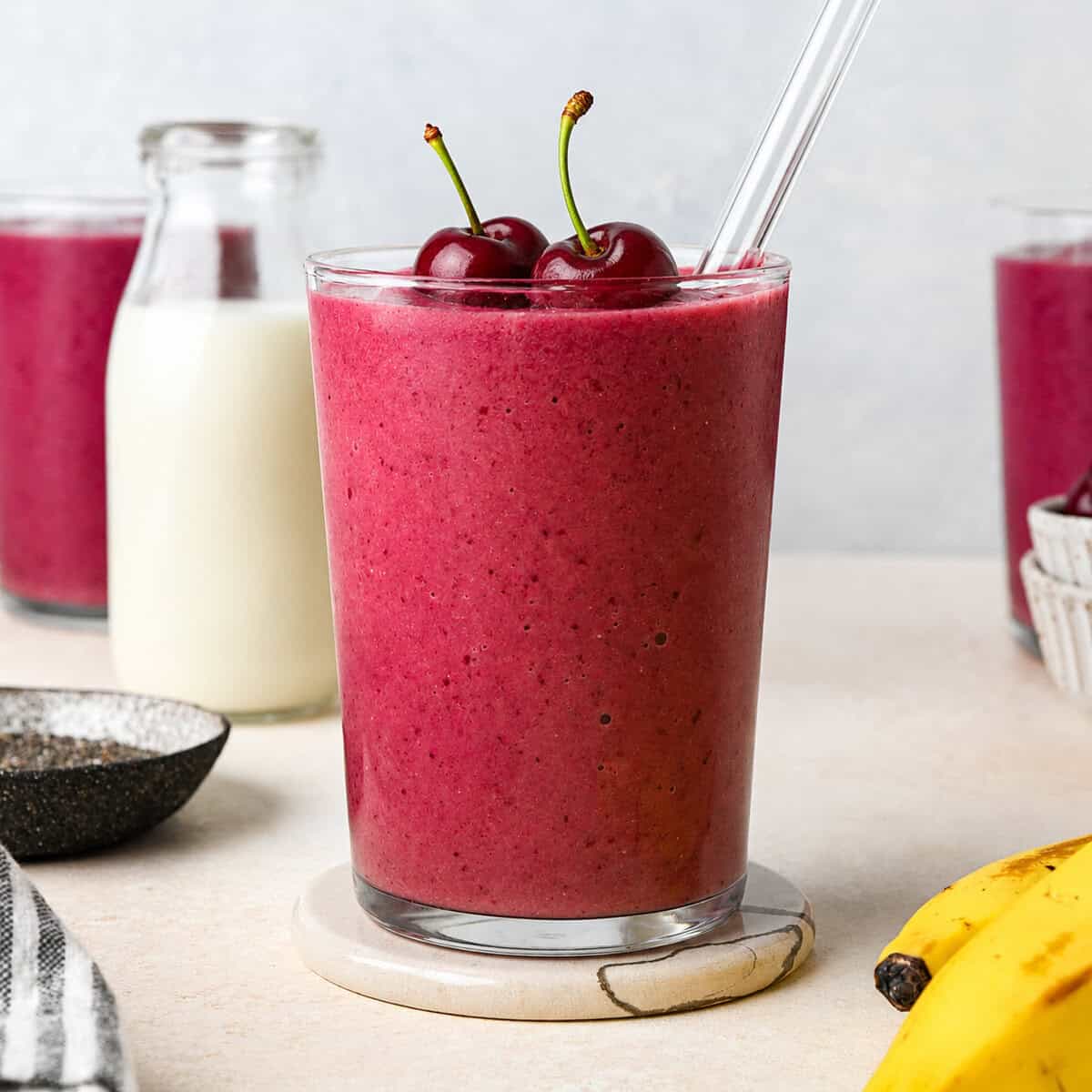 cherry smoothie recipe in a glass topped with two cherries and there's a glass straw in it. There are two other glasses of cherry fruit smoothie, a glass of milk, a bowl of chia seeds and a bowl of cherries in the background. There are two bananas in the foreground