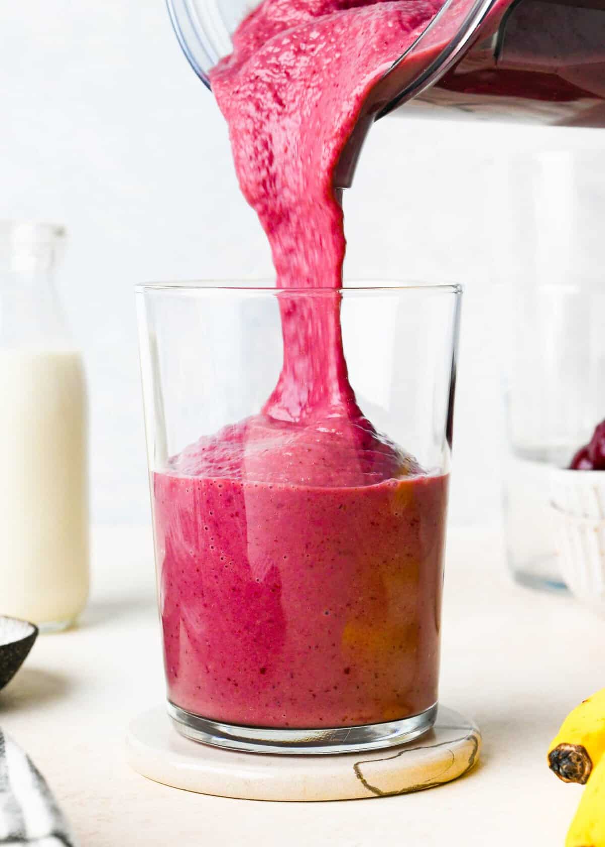 banana cherry smoothie recipe being poured from a blending container into a glass, it's thick texture is visible. there is a glass of milk, bowl of cherries and banana in the background.